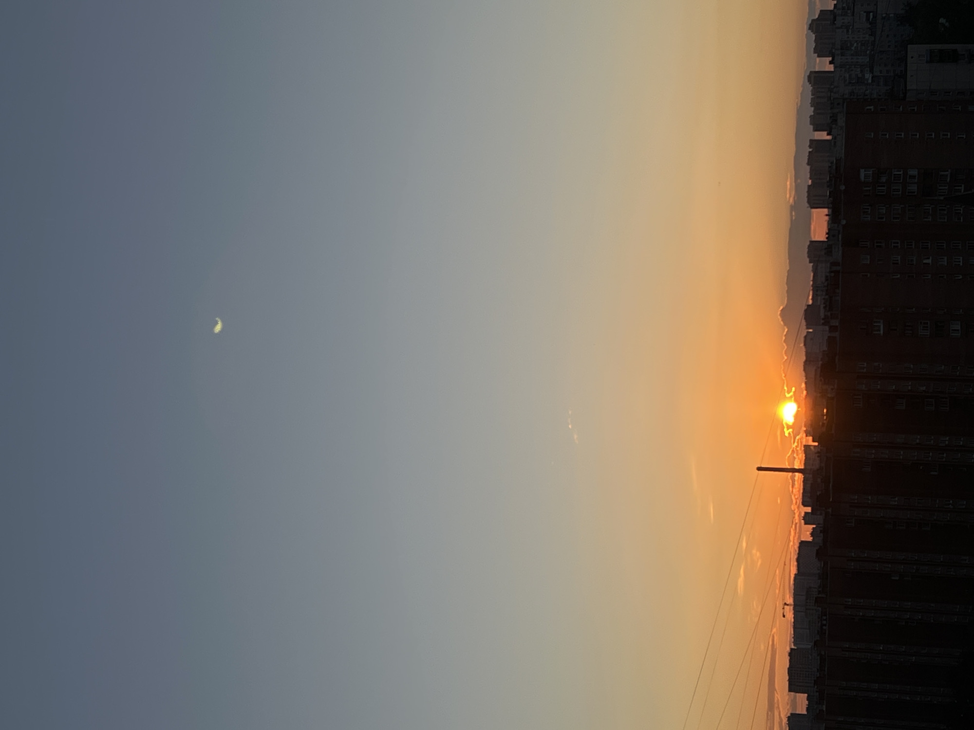 This is a vertically oriented photograph taken during sunset over a cityscape. The sky transitions from a soft, pale blue on the left to a warm, vibrant orange and yellow on the right, indicating the setting sun. In the upper left quadrant of the sky, a sliver of the moon is faintly visible.
The right side of the image is dominated by a silhouette of buildings. These appear to be apartment blocks, characterized by rows of windows and their imposing height. A tall, slender structure resembling a chimney or smokestack stands amongst the buildings. The setting sun is a bright, concentrated point of light near the horizon, casting a warm glow and creating a hazy effect around it, partially obscured by wispy clouds. The overall impression is one of a city at dusk, with the natural beauty of the sunset contrasting with the man-made urban environment. The image evokes a sense of calm and the end of a day. There is no visible text in the image.