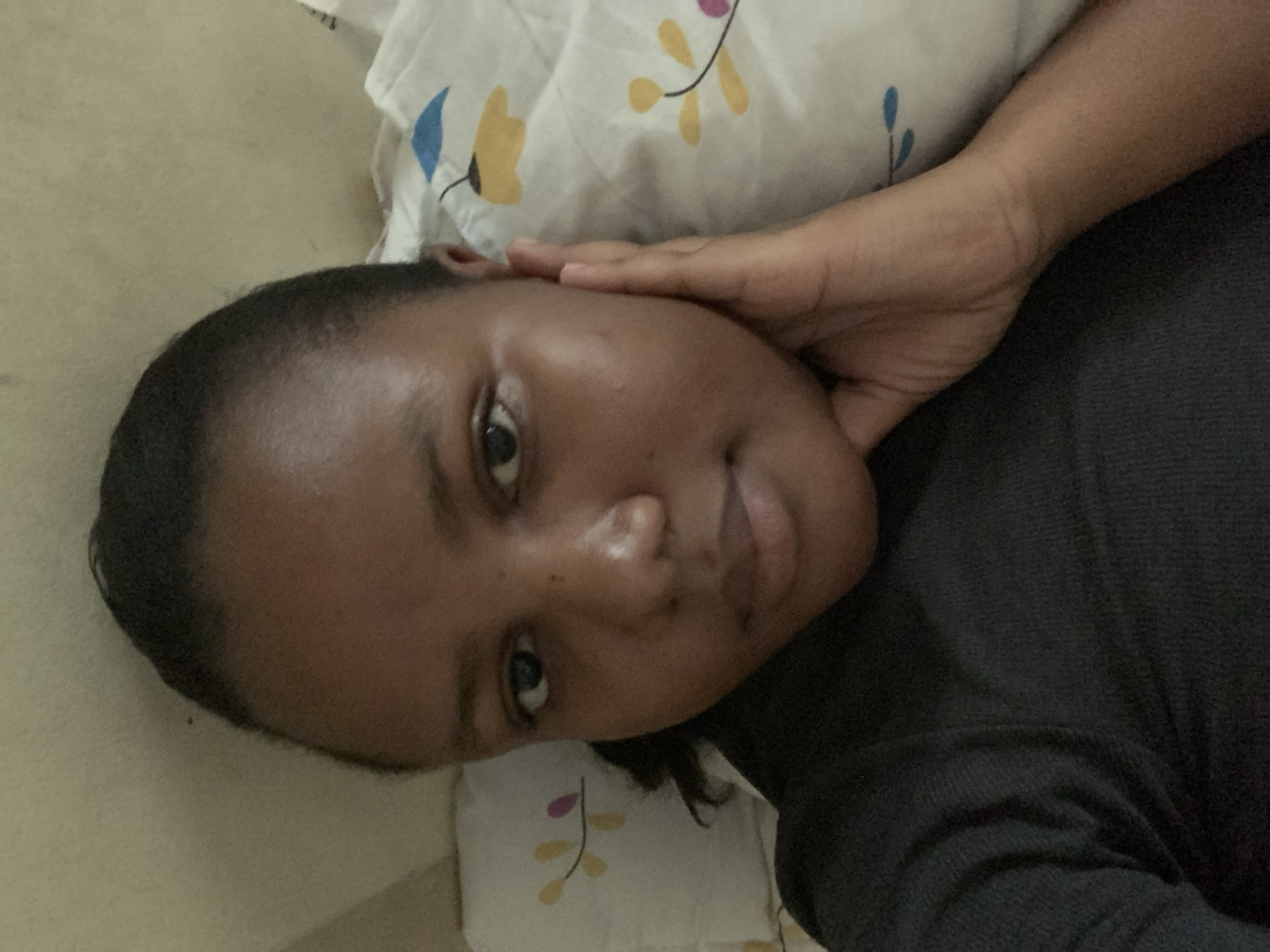 A close-up, horizontally oriented photograph captures a person lying down indoors, likely in a bedroom in Dar es-Salaam, Tanzania. The subject is an individual with dark skin and short, closely cropped dark hair, looking directly into the camera. Their head rests on a white pillow adorned with a subtle floral pattern featuring blue, yellow, and faint pink/purple leaf or flower motifs.
The person's right hand is placed gently against their temple or forehead area. They are wearing a dark-colored, possibly black or dark grey, short-sleeved garment, the texture of which suggests a soft fabric like cotton. The background consists of the patterned pillow to the right and a plain, light-colored surface, possibly a wall or headboard, to the left.
The individual's expression is neutral to slightly serene, with their dark eyes open and focused. There is a visible natural sheen on their forehead and the bridge of their nose, indicating either skin oils or the effect of the lighting. The overall impression is one of relaxation or a casual pose. No text is visible in the image.