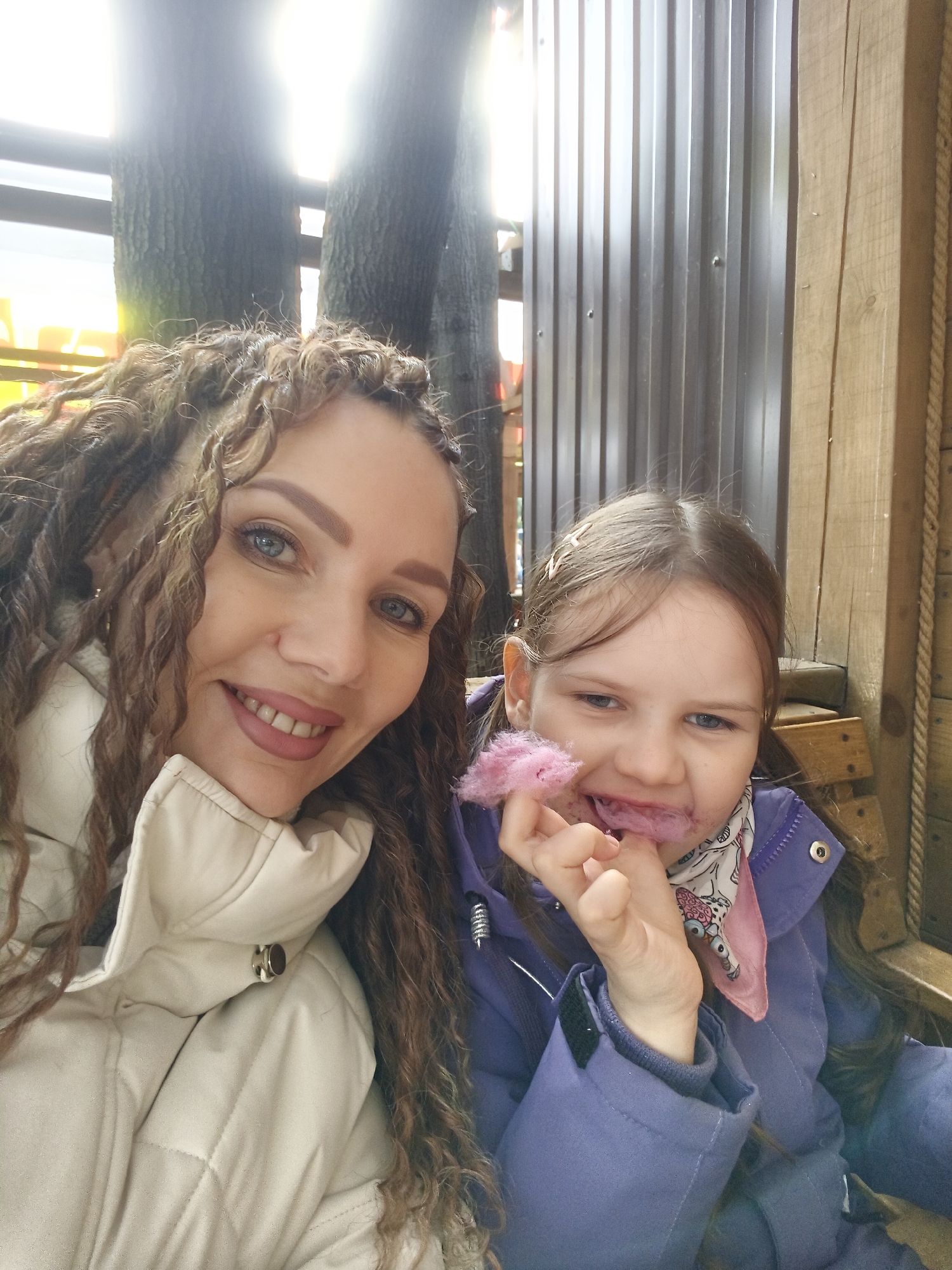 The image presents a woman and a young girl, likely in an outdoor setting. The woman is wearing a light-colored, quilted jacket and has long, curly brown hair. She is smiling at the camera. The girl, seated beside the woman, is wearing a blue jacket and appears to be eating cotton candy. There are metal and wooden structures in the background. Tree trunks are also visible behind the woman.