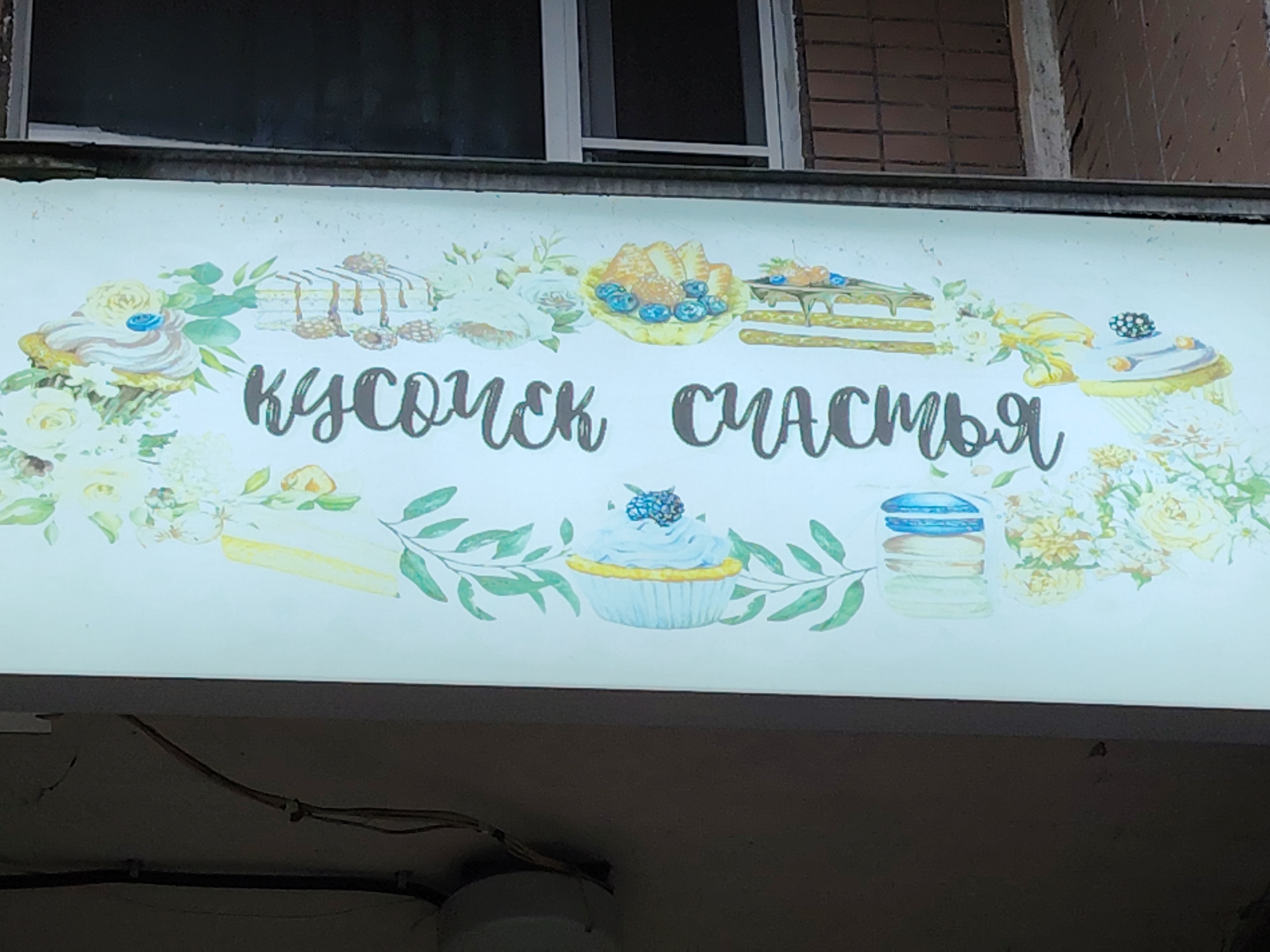 The image shows a lit sign with an illustration of various desserts, including cupcakes, macarons, and cake slices, decorated with floral elements. The text on the sign, written in Cyrillic script, reads "кусочек счастья," which translates to "a piece of happiness." This sign is likely for a bakery or dessert shop. The background shows a brick wall and a dark window, suggesting an outdoor setting. Based on the text and the style of the sign, this scene is likely located in Zelenograd, Russia.