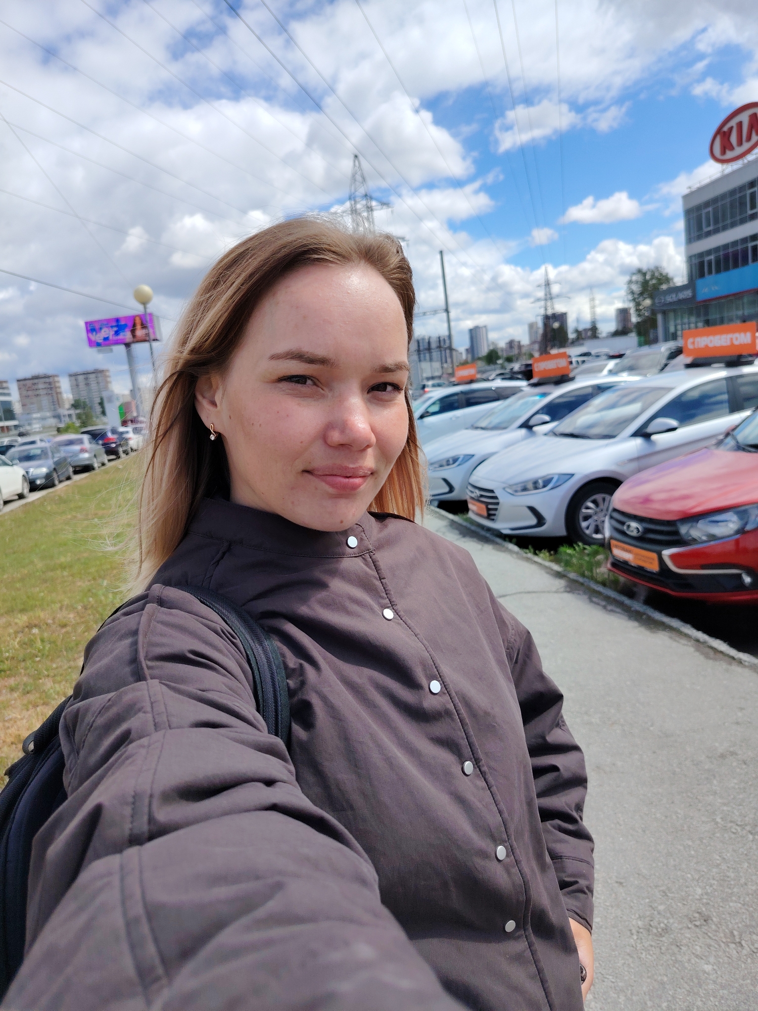 A young woman takes a selfie in Yekaterinburg, Russia. She is standing on a sidewalk next to a parking lot full of cars at a KIA dealership. She has shoulder-length blonde hair, fair skin, and is wearing a dark-colored button-down jacket and a backpack. The day is sunny with some clouds in the sky. In the background are several buildings, billboards, and power lines.