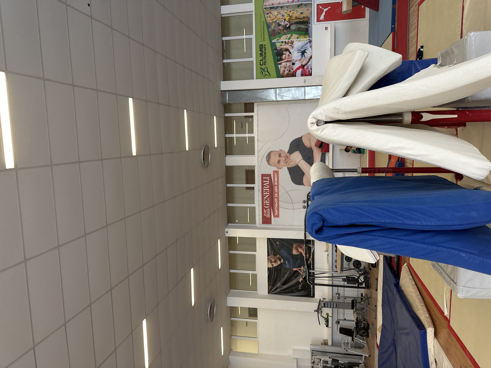 The image captures an indoor sports facility, likely a gymnasium or training center, with a brightly lit ceiling covered in rectangular acoustic tiles and long, linear LED light fixtures. A round clock is mounted on one of the walls.
Several large posters and banners decorate the walls. A prominent banner displays the "GENERALI" insurance logo with the Cyrillic slogan "ПАРТНЬОР ЗА ЦЯЛА ЖИВОТ" (Partner for a whole life), featuring a man in a black t-shirt. Above it, a green poster for "CLIMB ACADEMY" showcases images of children bouldering, alongside its logo and website "www.climbacademy.eu". A red "PUMA" logo is also visible nearby. Further down, another large, dark banner features a man in athletic wear, standing next to gym equipment.
The lower half of the walls features multiple windows, allowing some natural light into the space. The floor is covered with various exercise mats, including large blue and beige/white ones. Gym equipment, such as weight racks, benches, and exercise machines, are visible in the background and foreground. A folded white protective padding is draped over a red support bar, typical of gymnastics or athletic apparatus. No people are actively engaged in sports; only images of people are present on the banners. The overall impression is that of a modern, well-equipped training facility, situated in Sofia, Bulgaria.