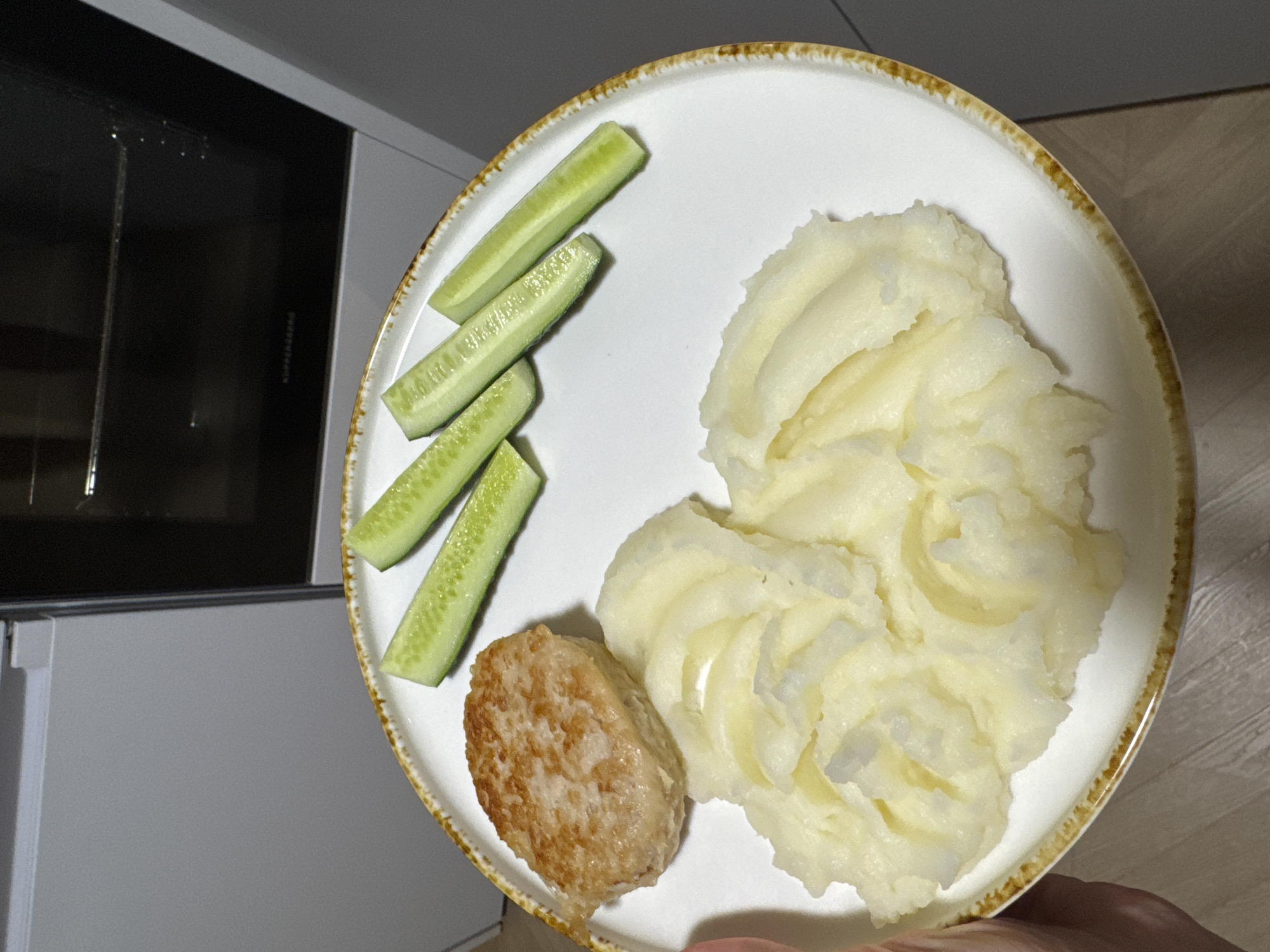 The image shows a plate of food on a table in front of a modern kitchen appliance, likely a built-in oven. The plate contains a single, round, golden-brown cutlet or burger patty, a generous serving of mashed potatoes, and four sliced pieces of cucumber arranged in a line. The plate has a white surface with a textured, yellowish-brown rim. The background is blurred but appears to be a kitchen environment with a refrigerator and the aforementioned oven. The food is presented in a home setting. The image was captured in Postol, Russia.