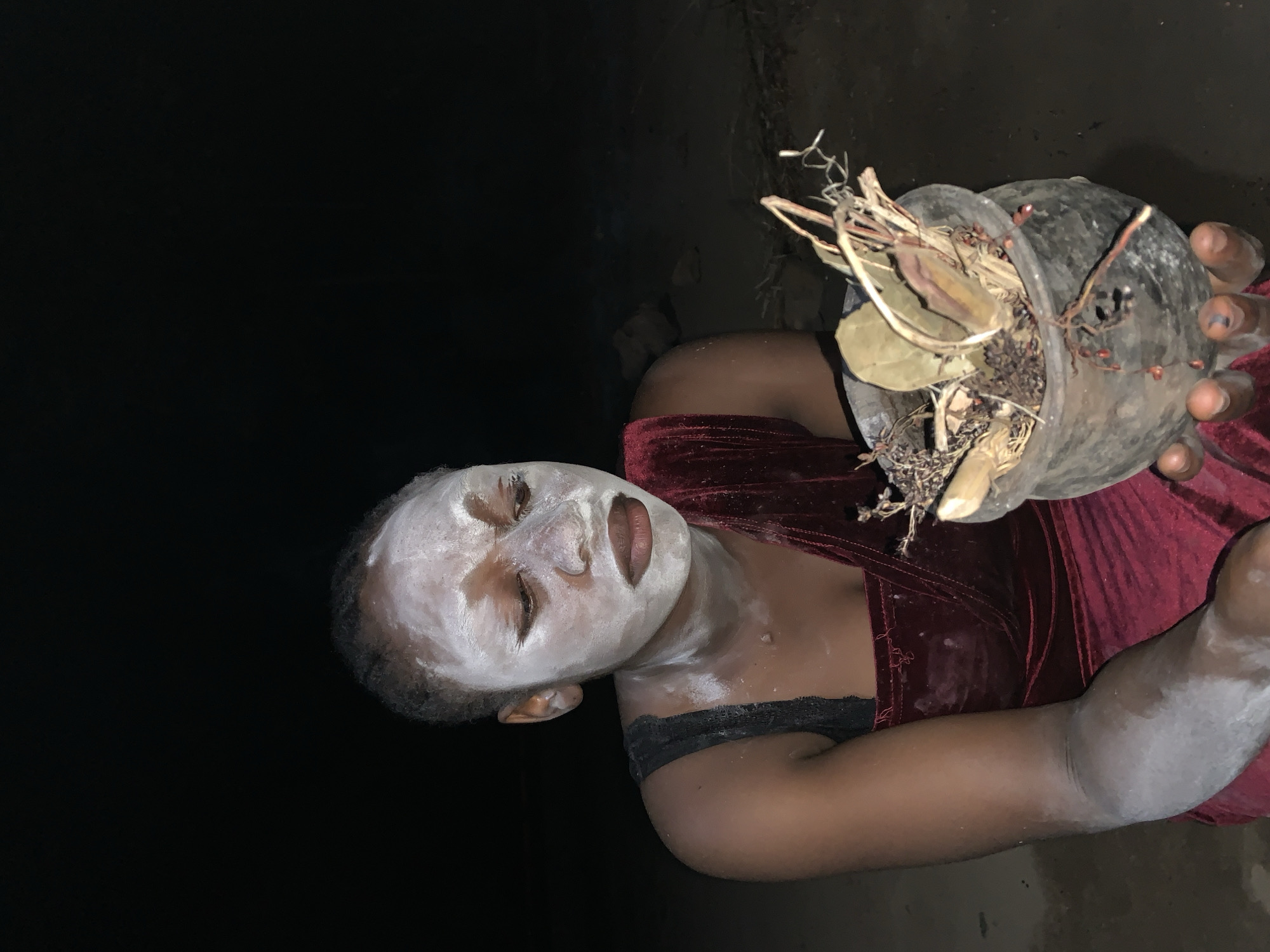 In a dimly lit environment, a dark-skinned person with white facial markings holds a dark-colored container filled with dried leaves, twigs, and roots. The person is wearing a dark red velvet top with black straps visible. The person's face, neck, and arm are smeared with white. The overall setting is dark with little ambient light. It is not possible to determine if the location is in the city of Agban, Nigeria.