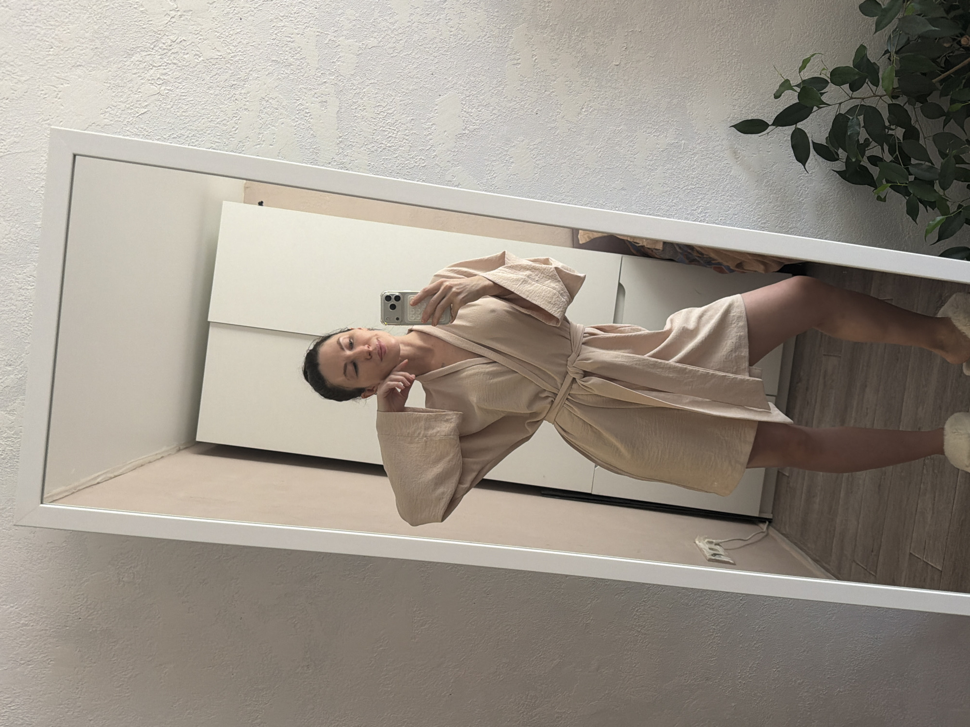 A woman is reflected in a mirror. The woman is wearing a light beige robe. She is holding a phone up to take a picture of herself in the mirror. The background appears to be a room with a white wall and a plant in the upper right corner of the mirror's reflection. The floor appears to be light wood. The location is not identifiable as being in Perm, Russia.