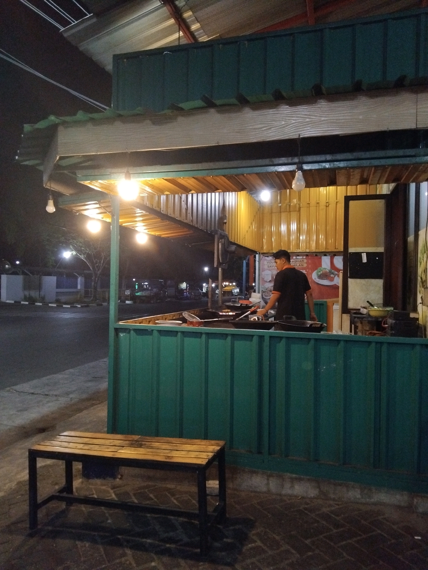 An outdoor food stall with a green metal facade is illuminated by warm lights at night in the city of Kepuh Kiriman, Indonesia. A man wearing a black t-shirt and dark pants stands behind the counter, preparing food. He is facing away from the camera and appears to be cooking. In front of the stall, a wooden bench with a dark metal frame sits on a brick-paved area. The street is visible to the left, with a few parked vehicles and trees in the background. The overall scene suggests a typical evening setting for a street food vendor.