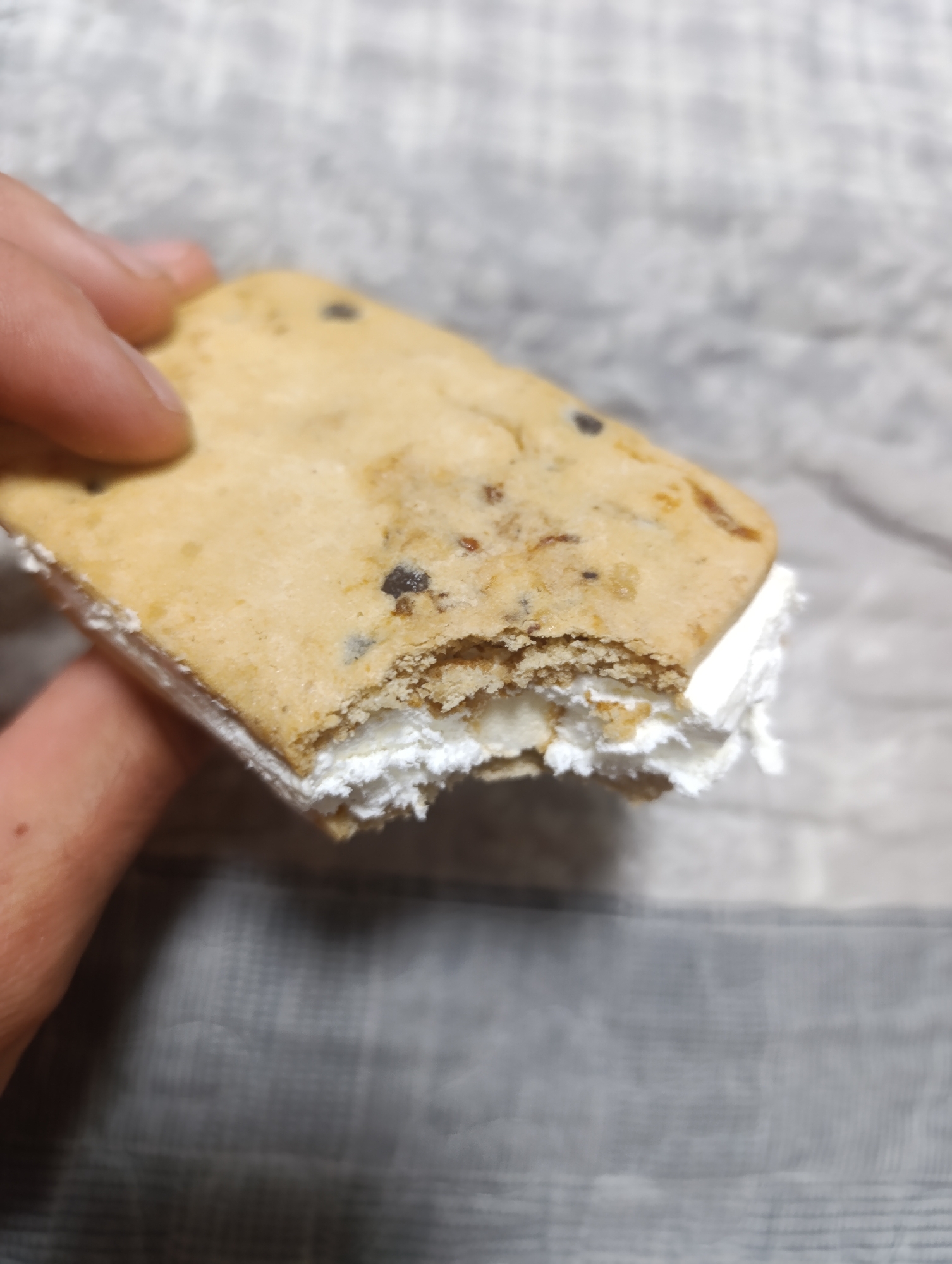A close-up, slightly angled shot shows a person's hand holding a chocolate chip cookie ice cream sandwich. The cookie is golden brown and appears to be slightly crumbly, with visible dark chocolate chips scattered throughout. The sandwich filling is a thick, white, creamy substance, likely ice cream or marshmallow creme, which is exposed on the bitten edge, revealing its fluffy texture. The background is out of focus, showing a mottled grey surface that could be a table or countertop, with a texture reminiscent of stone or a textured fabric. The lighting is bright, highlighting the textures of the cookie and the filling. The image does not contain any information indicating a location in Zlatopil, Ukraine.