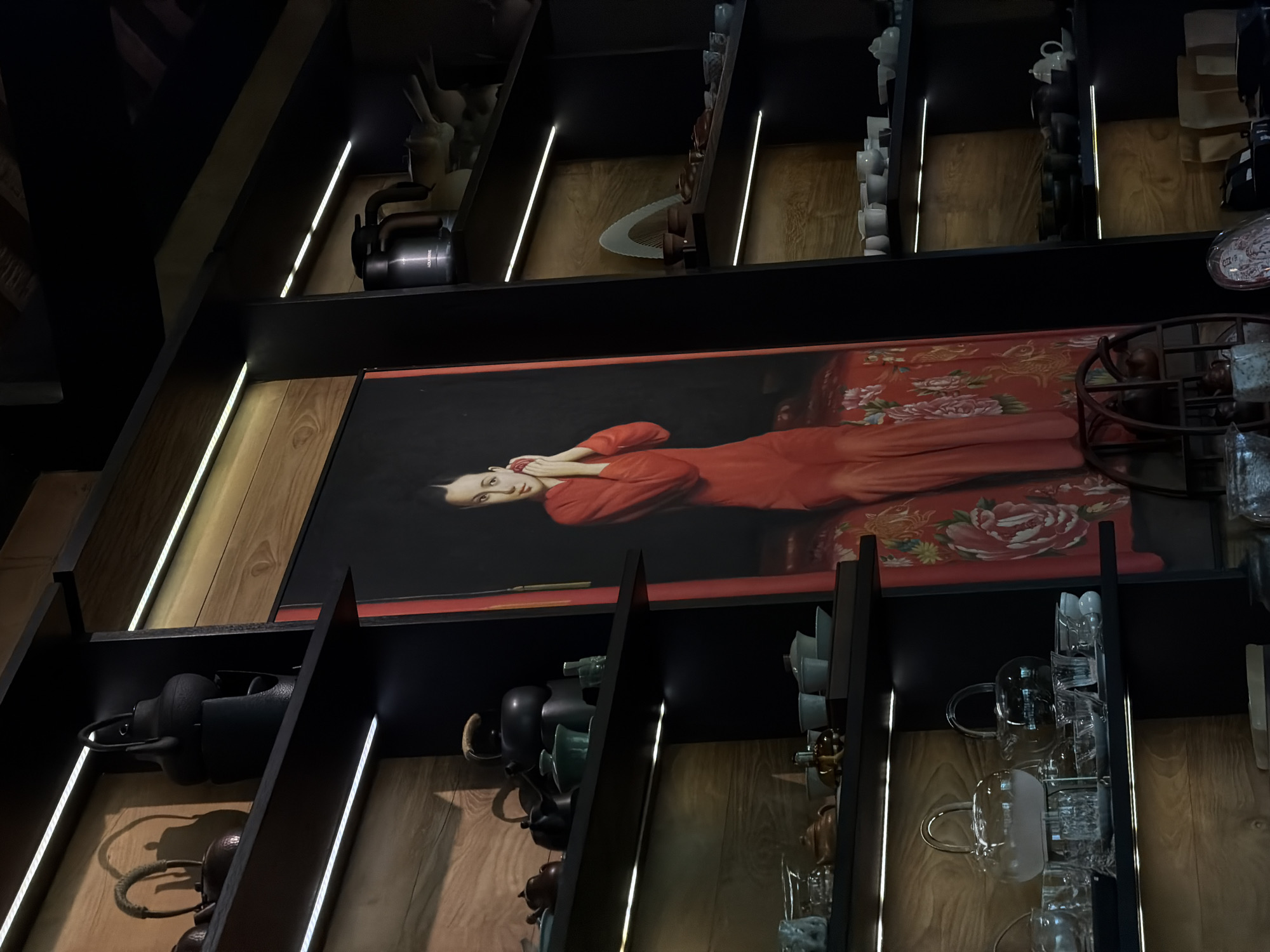 The image displays an interior retail or gallery space in Singakerta, Indonesia, characterized by prominent display shelving and a large portrait. A vertically oriented painting, featuring an East Asian woman in a red traditional dress, seated on a richly patterned red fabric against a dark background, is centrally positioned. Her dark hair is pulled back, and her hands are clasped near her face. The painting is flanked by dark, multi-tiered display shelves with light-colored wooden back panels.
Each shelf level is illuminated by horizontal LED strip lights. The shelves on the left exhibit various teaware, including several dark, possibly cast-iron, teapots or kettles on the upper levels. Below them, an assortment of smaller ceramic pieces in dark and lighter green/blue tones, alongside clear glass teapots, cups, and bowls, are arranged.
The shelves on the right display dark cylindrical containers, one visibly labeled "AQUA STONE," a white fan-shaped decorative object, and numerous white and dark ceramic cups, bowls, and saucers. Brown paper bags or boxes are present on the lowest visible shelf section. A dark, circular, multi-tiered wooden display stand is partially visible in the lower-center, overlapping the base of the painting. No individuals are present in the scene, and no actions or interactions are depicted.
