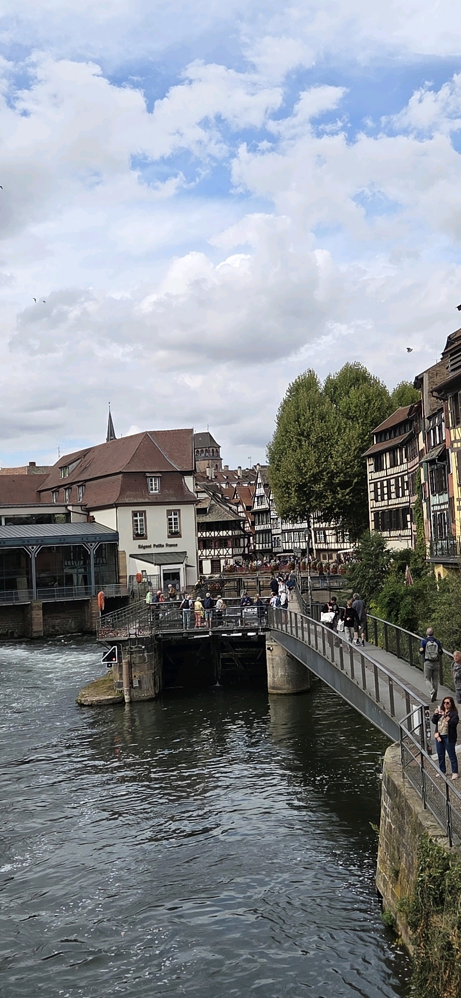 The image depicts a view of the Petite France district in Strasbourg, France, with a river and a bridge. The river is dark and reflective, with some ripples and white foam. A metal pedestrian bridge spans the river, with several people walking across it. On the left side of the bridge, there are buildings with red roofs and traditional French architecture, some featuring half-timbered structures. On the right side, more buildings with similar architectural styles are visible, some with balconies and window boxes. The sky is partly cloudy with patches of blue. On the bridge, a woman in a dark jacket and jeans is visible, holding up a camera to take a picture. Another person, a man with a backpack, is walking further across the bridge. A group of people are gathered on a lower platform to the left of the bridge, near a building with signage that reads "Régent Petite France." The overall scene is one of a bustling tourist area with historical architecture and a scenic riverfront.