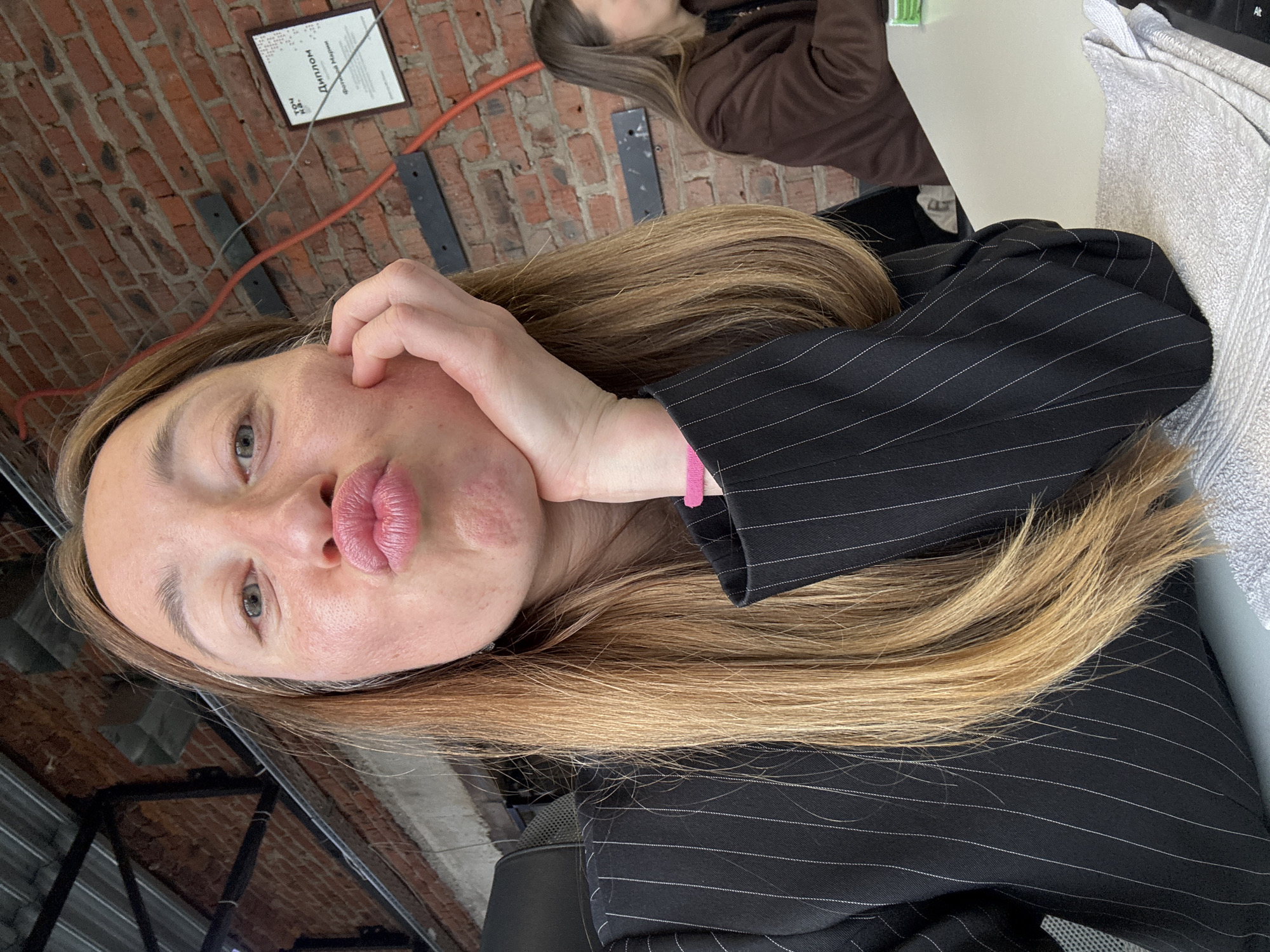 A fair-skinned woman with blonde hair poses indoors. She wears a black pinstripe blazer and rests her hand on her face, puckering her lips. Behind her is a brick wall with metal supports and a framed document in Russian. The background includes another person with brown hair and a brown top, a white table, a white towel, and a keyboard. The image was taken in Chaikovsky, Russia, as evidenced by the Russian text in the framed document.