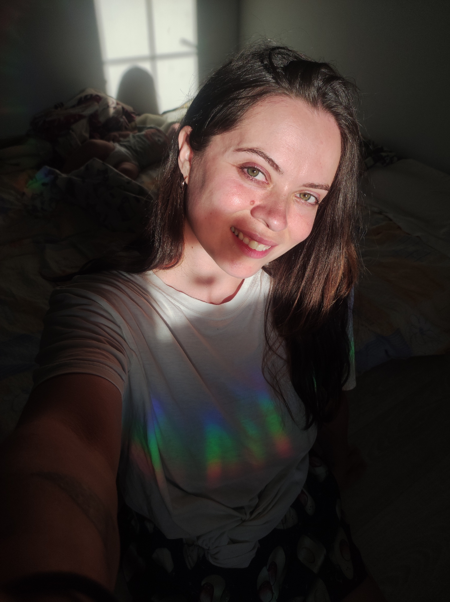 A Caucasian female, appearing to be an adult, is captured in a selfie in an indoor setting, presumably a bedroom, in the city of Unknown, Unknown. She has long, dark brown hair and light green eyes. Her face is illuminated by natural light from the right, highlighting a mole superior to her right nostril. She is smiling, revealing her teeth, and looks directly at the camera. She wears a white short-sleeved t-shirt, tied in a knot at the bottom, and dark shorts patterned with what appear to be light-colored flip-flops. A distinct rainbow spectrum, originating from a light source, is projected horizontally across her white t-shirt. In the background to her left, a bed with multiple blankets is visible. On this bed, a sleeping infant or small child is partially discernible under the covers. A window frame's shadow, along with a distinct, rounded shadow of a head or object, is cast onto the wall behind her, indicating strong natural light. The overall lighting is high-contrast, with parts of the scene in bright illumination and others in deep shadow.