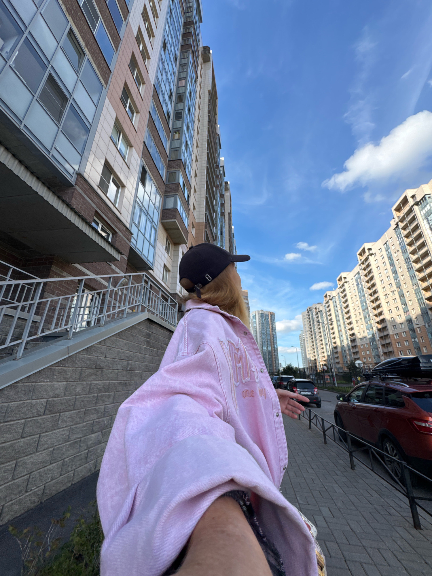 The image depicts a person wearing a pink jacket and a black baseball cap, facing away from the camera and looking up at a blue sky with scattered clouds. The person is standing on a paved sidewalk next to a brick wall and a set of stairs leading up to a building entrance. In the background, tall, modern apartment buildings line a street with cars parked along the side. The scene appears to be an urban environment in Ladozhskoe Ozero, Russia. The sunlight suggests it is daytime, likely late afternoon or early evening given the angle of the light.