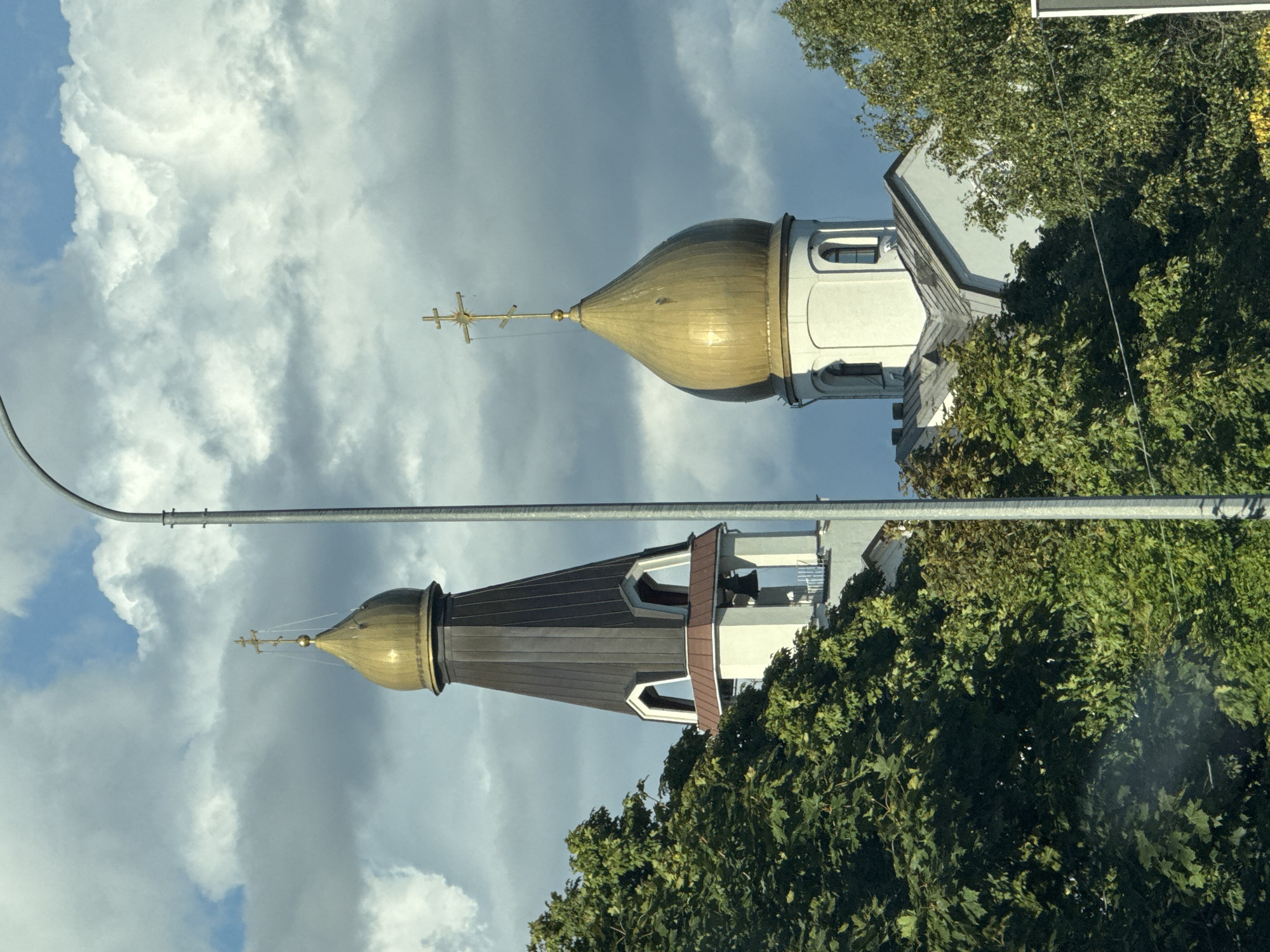 The image shows a close-up view of two onion domes topped with crosses, characteristic of Russian Orthodox churches, against a dramatic sky with heavy, grey clouds interspersed with patches of blue. A tall, slender grey lamppost runs vertically through the center of the frame, partially obscuring the church structures. Lush green trees frame the lower portion of the image, suggesting an outdoor setting. The larger, darker dome structure appears to be a bell tower, with arched openings revealing a bell. The smaller, golden dome is round and topped with a multi-tiered cross. The architecture suggests a religious building in a temperate climate, likely during daytime. The presence of the lamppost and the framing trees implies the photo was taken from a street or roadside. This structure is likely the Church of the Resurrection in Sestroretsk, Russia, which features similar golden domes and a bell tower.