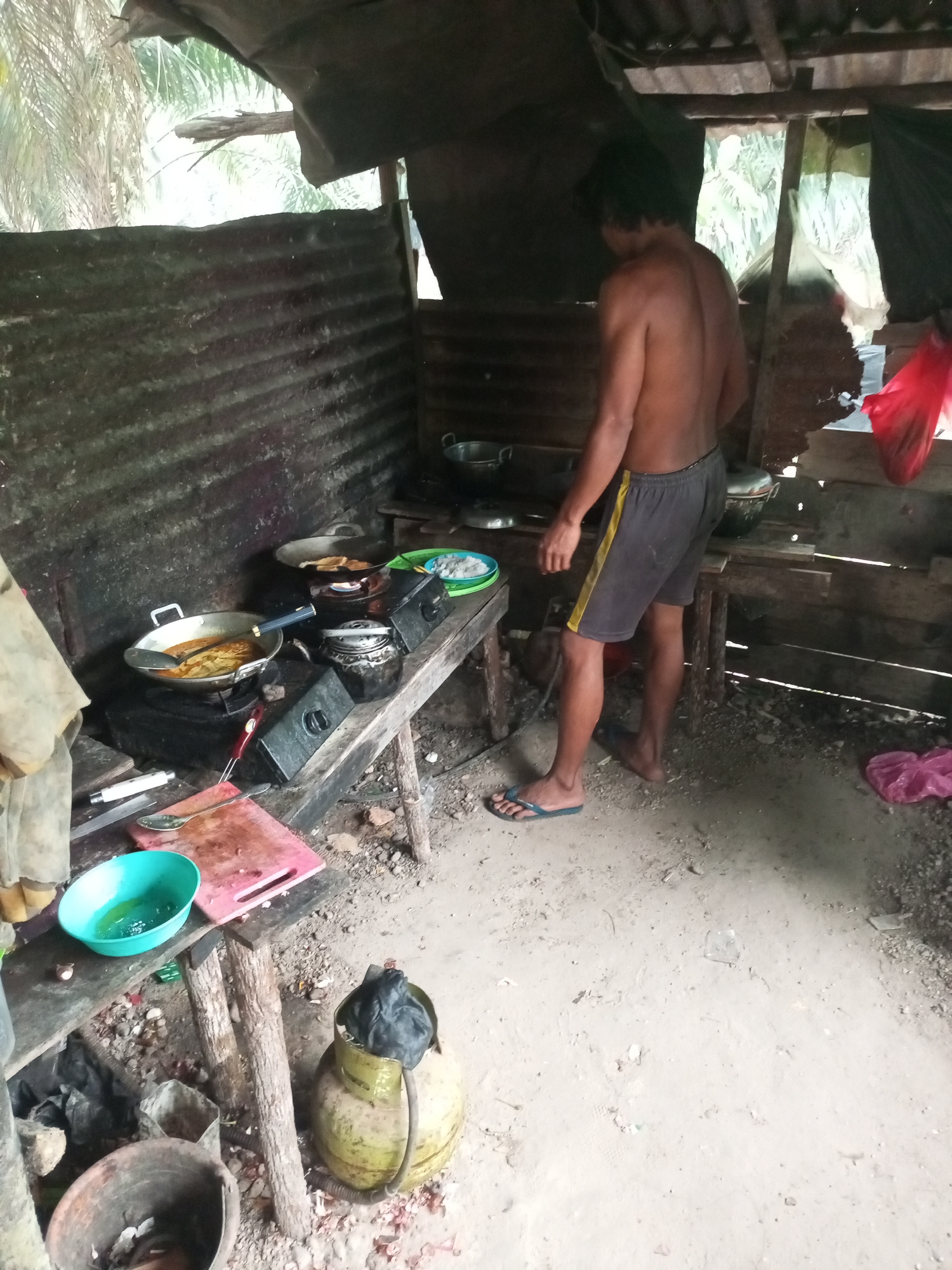 In a rustic, open-air kitchen environment, a shirtless man with dark hair stands facing away from the camera. He is wearing dark shorts with a yellow stripe and blue flip-flops. The kitchen appears to be constructed from corrugated metal and wood. A wooden table holds various cooking utensils and ingredients. On the table, two gas stoves are visible. The left stove has a frying pan with food cooking in it, and the right stove has a pot. A green bowl containing liquid sits on the table, next to a pink cutting board. A gas cylinder is placed on the ground near the table, with a black bag on top. The floor of the kitchen is dirt and sand, with some debris scattered around. In the background, foliage from palm trees is visible through gaps in the structure. The scene suggests a basic, functional cooking setup, possibly in a rural area, which aligns with the general description of Keling Panggau, Indonesia, as a village or rural community.
