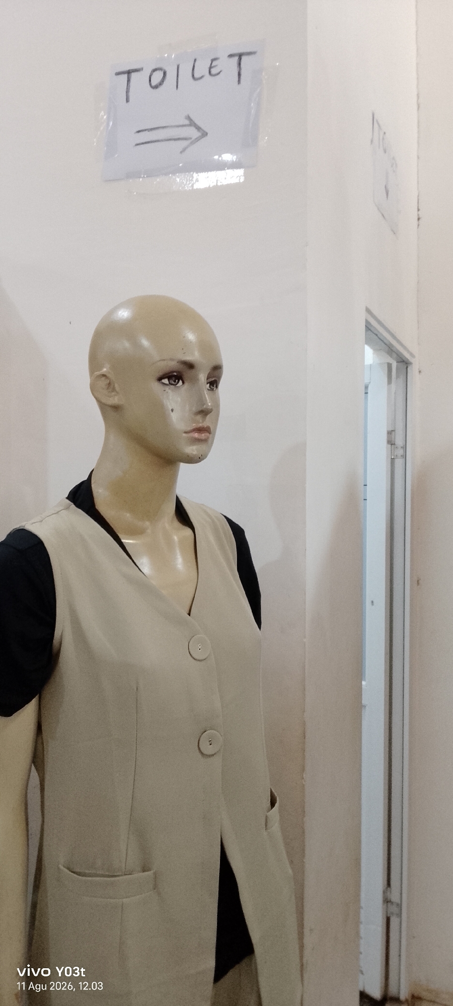 A beige mannequin wearing a light beige vest over a black shirt stands in an indoor setting. A sign on the wall above and to the left of the mannequin reads "TOILET" with an arrow pointing to the right. To the right of the mannequin is a doorway with a white frame and what appears to be a partially open door. The walls are painted a light color. A timestamp in the lower left corner reads "vivo YO3t 11 Agu 2026, 12.03". There is no indication of the city of Atula, Indonesia.