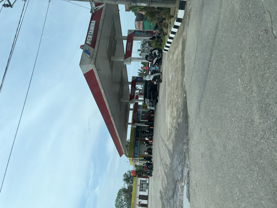A Pertamina gas station stands under a cloudy sky. Motorcycles cluster near the pumps, some with drivers, others parked. The station's canopy has a red and white trim. Adjacent is a building with yellow accents. A black and white striped curb runs along the side of the road, which is cracked. Utility poles and wires are visible in the sky.