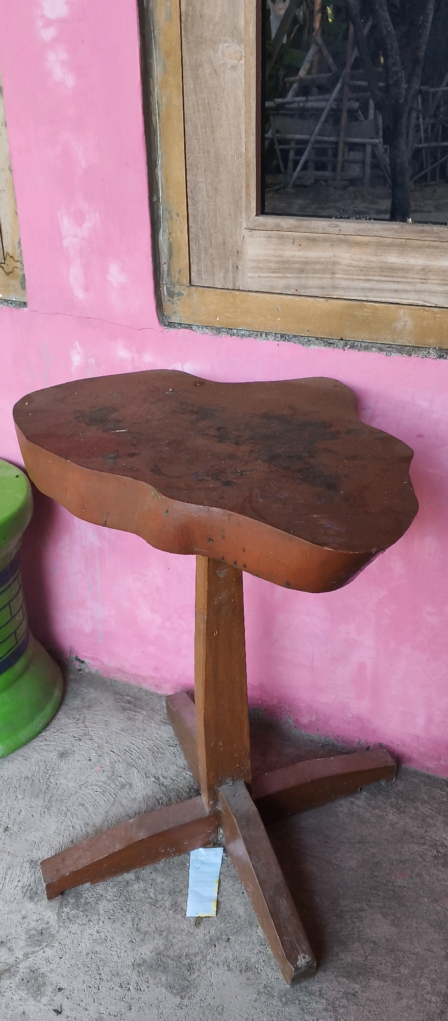 A small, irregularly shaped wooden table with a dark brown, possibly lacquered, top stands on a concrete surface against a pink wall. The table has a single, thick wooden pedestal that splits into four legs forming a cross-shaped base. A small piece of torn paper, possibly a receipt or label, is affixed to the concrete near the base of the table. To the left of the table, a portion of a green and purple cylindrical object, possibly a stool, is visible. Behind the table, a window with a wooden frame is set into the pink wall, and through the window, a reflection of scaffolding and trees can be seen. The scene appears to be outdoors, given the natural light and outdoor elements. The location is specified as Japanan, Indonesia.
