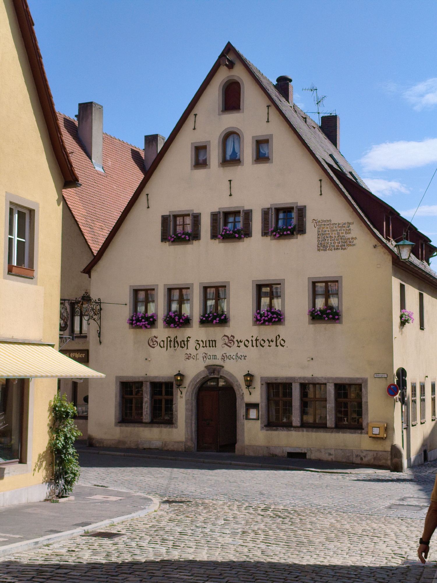 This image shows a quaint cobblestone street scene in Rothenburg ob der Tauber, Germany, featuring a traditional gabled building with a sign reading "Gasthof zum Broilerle." The building, painted a light yellow, has multiple windows adorned with colorful flowers, and dark wooden shutters. A dark arched doorway is visible on the ground floor, and a decorative wrought-iron sign hangs from the facade. The sky above is a clear blue with a few wispy clouds. To the left, a portion of a yellow building with a striped awning is visible, and a green plant grows up the side of it. The street itself is paved with rounded cobblestones, and a portion of a person's arm and hand is visible in the lower right corner of the frame, suggesting they are walking away from the camera.