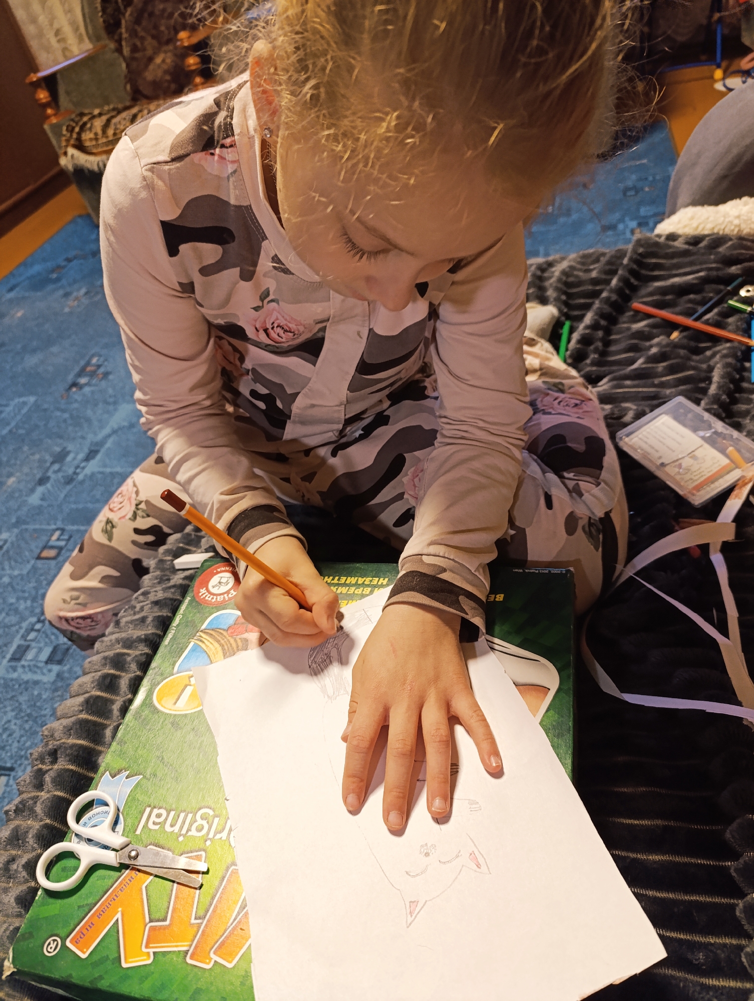 A young girl is drawing a cat on a piece of paper. She is sitting on the floor on a blanket, and there are colored pencils, scissors, and a game box nearby. She is holding a pencil in her right hand and using it to draw the cat's face. The location is Unknown, Unknown.