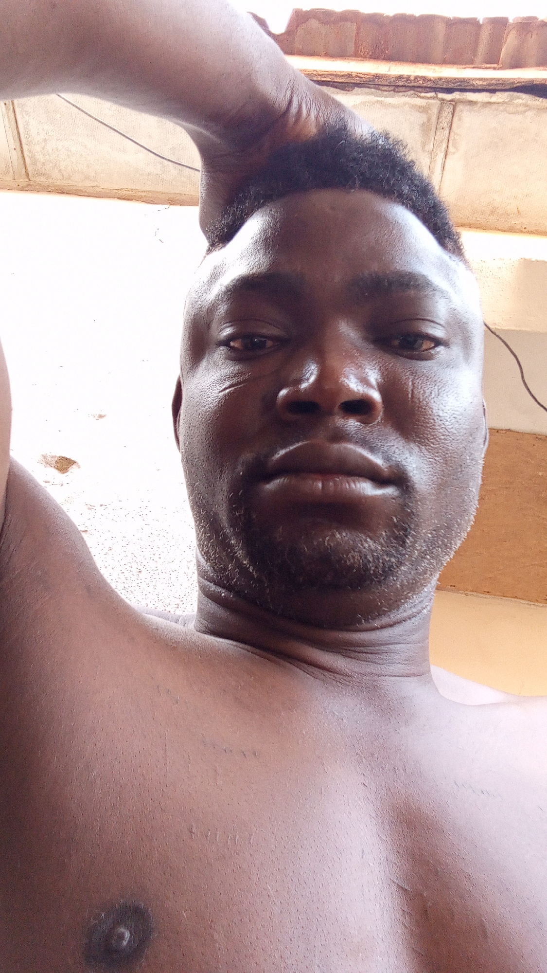 A close-up, low-angle selfie shows a dark-skinned man with a closely cropped black hairstyle and a short beard. The man has a solemn expression and is shirtless. His right arm is raised, with his hand near the top of his head. The background features a white wall with visible damage, possibly cracks or peeling paint, and a portion of a brown tiled roof is visible at the top of the frame. A wire runs across the top right corner. The location is Unknown, Unknown.