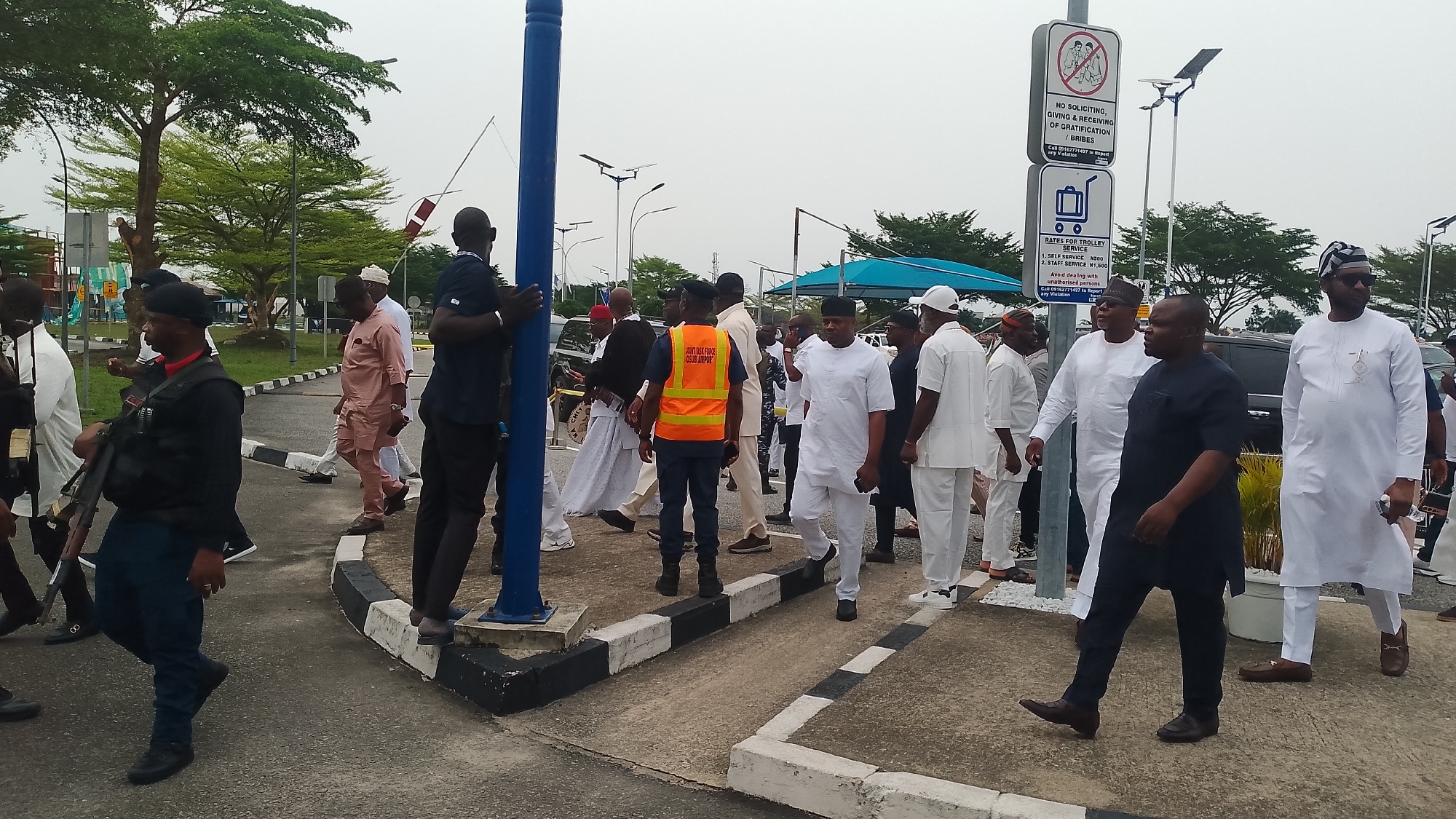 The image depicts a group of people in what appears to be an airport environment in Orerokpe, Nigeria, as indicated by the "OSUB AIRPOR" on the vest worn by a man in an orange vest. The group is diverse in dress, with some wearing traditional Nigerian attire, others in suits, and some in casual wear. At least one armed security personnel is present. A sign warns against soliciting, giving, or receiving bribes, and another lists rates for trolley service. The environment includes trees and buildings in the background.