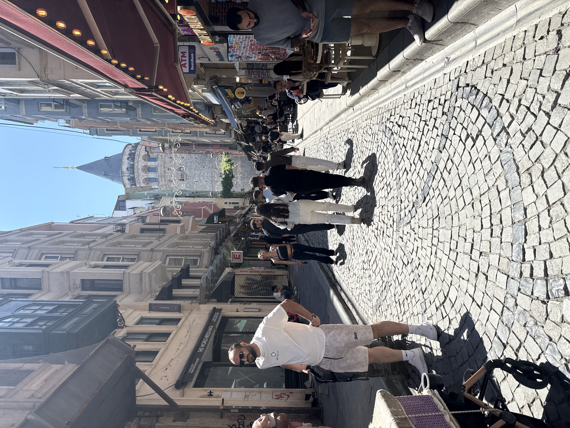The image shows a street view in Beyoğlu, Turkey, with the Galata Tower visible in the distance. The foreground features a cobblestone street with pedestrians walking, and a man in a white shirt and light-colored shorts standing on the right. A stroller is next to him. Buildings line both sides of the street, with shops and restaurants visible. Some establishments have awnings and outdoor seating. The sky is clear. Street signs and ATM signage are also visible.