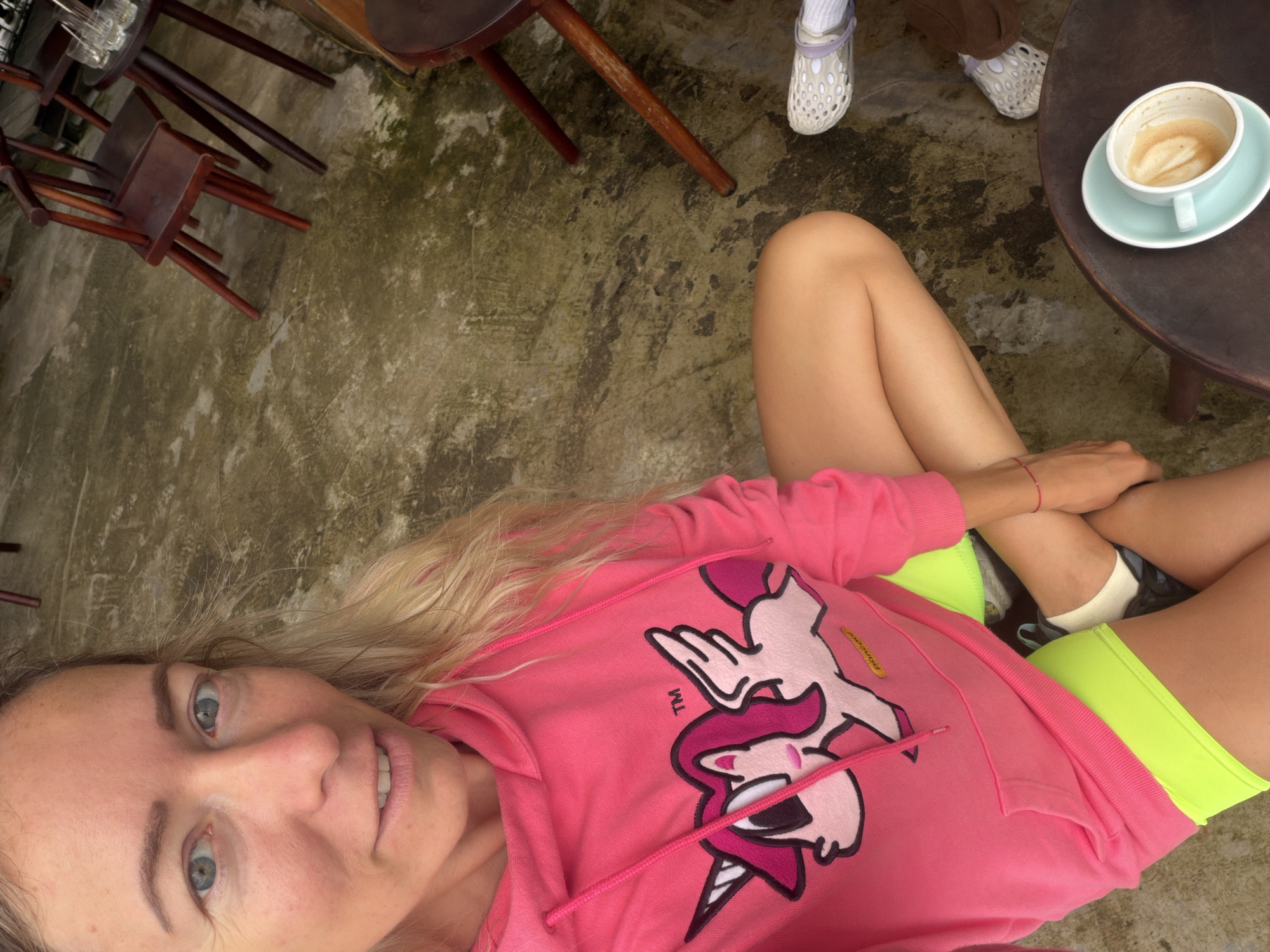 A woman with blonde hair is sitting cross-legged on a dirty, uneven outdoor patio. She is wearing a bright pink hooded sweatshirt with a cartoon unicorn design and neon green shorts. Her left hand is resting on her knee, and her right arm is bent with her hand near her face. Next to her, also seated, is a second person wearing white perforated shoes and brown pants, partially visible. A small, round, dark brown wooden table sits in the foreground with a white cup of coffee and saucer on it. In the background, there are several dark wooden chairs and tables. The ground is a mix of concrete and dirt, with patches of moss or algae. The overall scene appears to be a casual outdoor seating area at a cafe in Da Lat, Vietnam.
