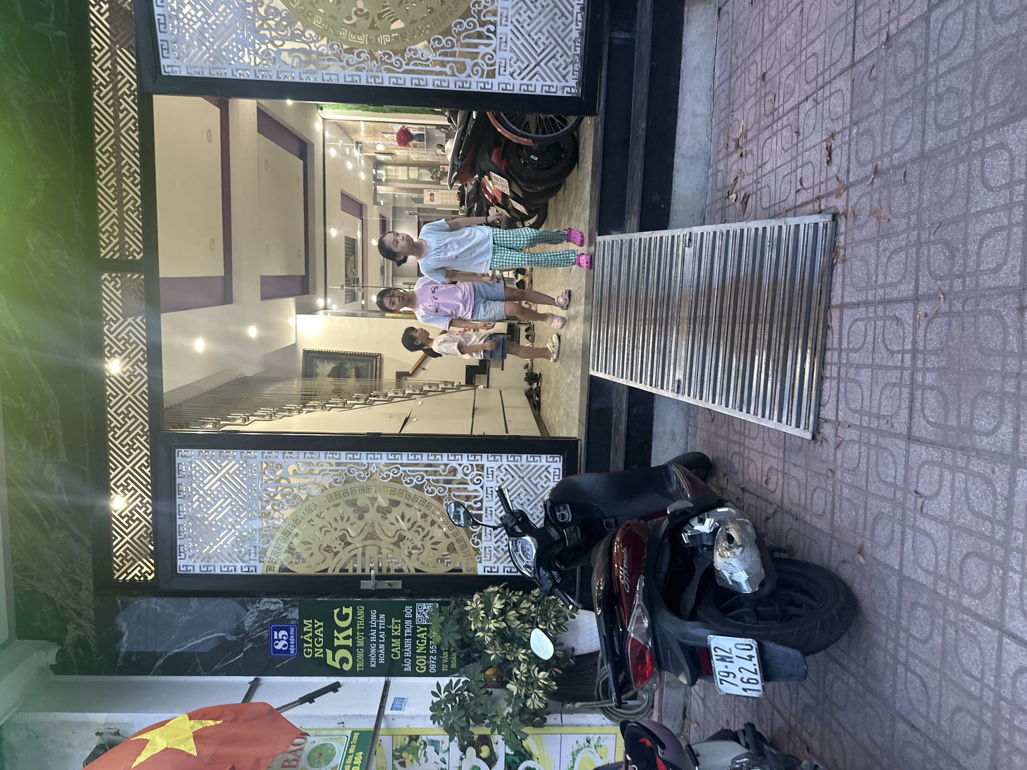 The image captures three children standing in the doorway of a building with an ornate, laser-cut metal door and a grated ramp in front. Inside, a staircase is visible. Two parked motorbikes are near the entrance. Signage with Vietnamese text is displayed, advertising discounts and services, including the phone number 0972 557. A Vietnamese flag is partially visible. The license plate of one of the motorbikes reads "79-N2 162.40". Based on information extracted from the signs, the location is possibly in the Điện Biên Phủ area of Nha Trang, Vietnam.
