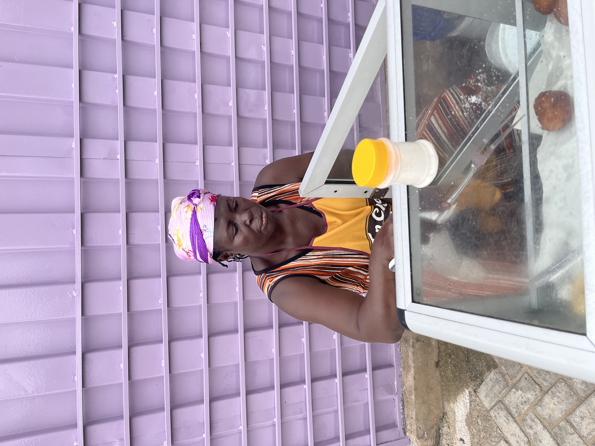 The image features a woman wearing a yellow and orange striped shirt and a patterned headscarf, positioned in front of a purple corrugated metal wall. She is standing next to a glass display case, through which several food items appear to be visible. Among these items are round, brown pastries, likely fried dough balls, and white powders. A yellow-capped container is also present near the edge of the display case. The scene suggests a commercial setting, possibly a street vendor or small shop, in Accra, Ghana. The lighting appears to be natural daylight. The woman's eyes are closed, and her mouth is slightly open, suggesting she might be resting or in the process of preparing or serving food. The overall atmosphere is that of an everyday scene, likely during the day.