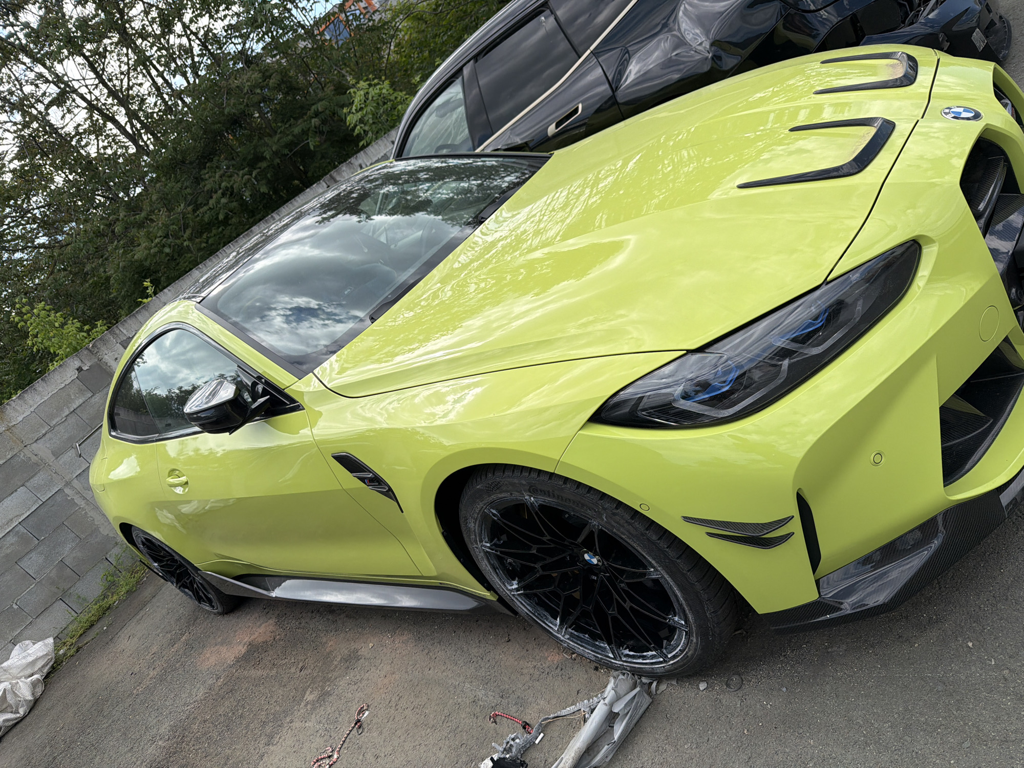 A vibrant, yellow BMW M4 is parked on a paved surface. Another, black vehicle is partially visible behind it. The location appears to be outdoors, with trees and a stone wall visible in the background, possibly in Burgas, Bulgaria. The BMW is the primary subject, featuring black rims and aerodynamic elements. The environment suggests a residential or industrial area. Debris is scattered on the ground near the front tire of the yellow car.