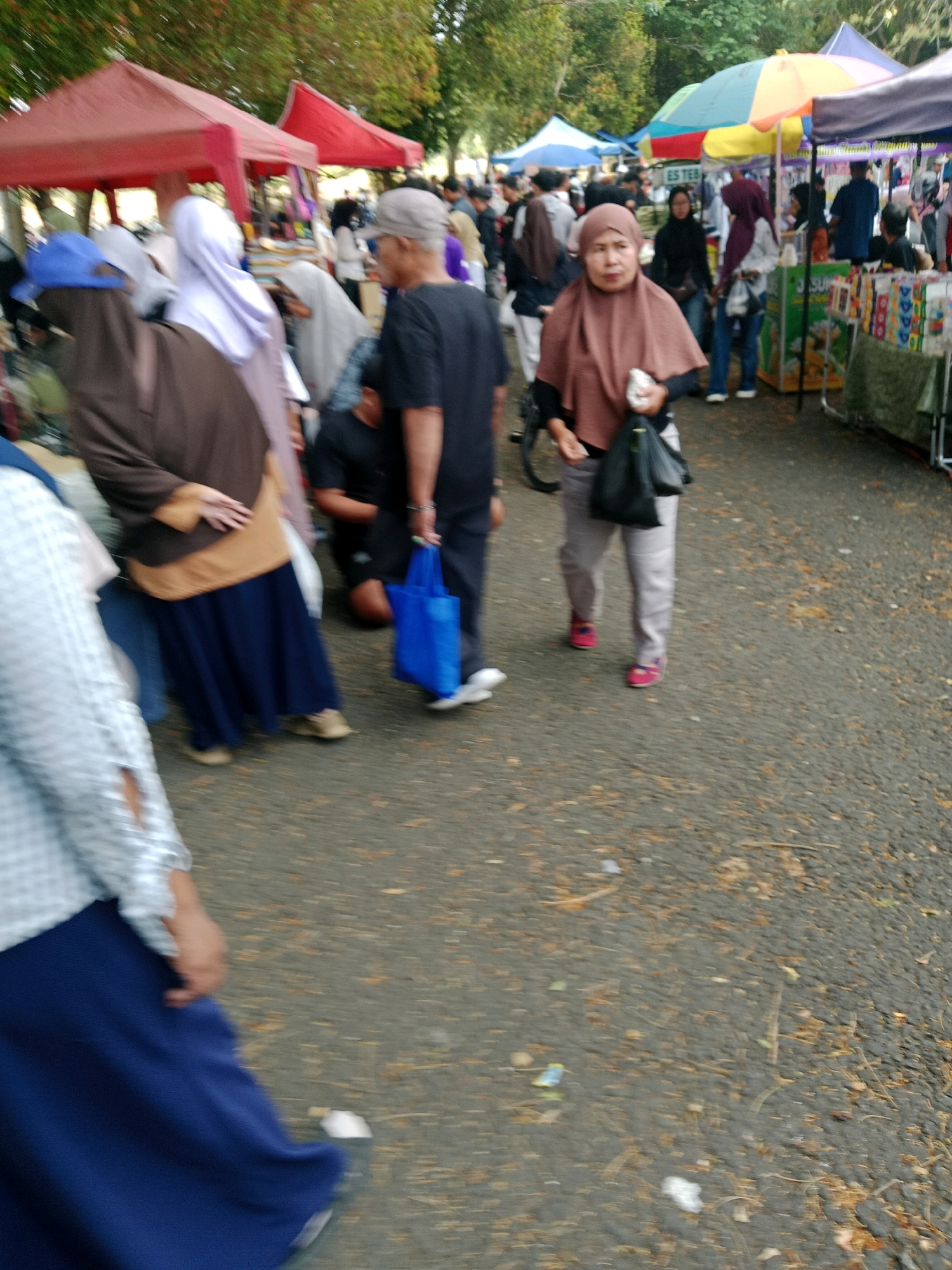 The image depicts a street scene, likely an outdoor market or festival, in the city of Ciamis, Indonesia. The foreground is dominated by a person wearing a blue headscarf and a brown garment, partially obscured. To their right, a person in a brown hijab and pink shoes is walking, carrying a black bag. Behind them, a person in a black shirt and cap is also walking. In the background, several market stalls with colorful canopies are visible, and many people are milling about. The ground is paved and scattered with leaves and debris. The atmosphere appears to be bustling, with a variety of goods being displayed at the stalls.