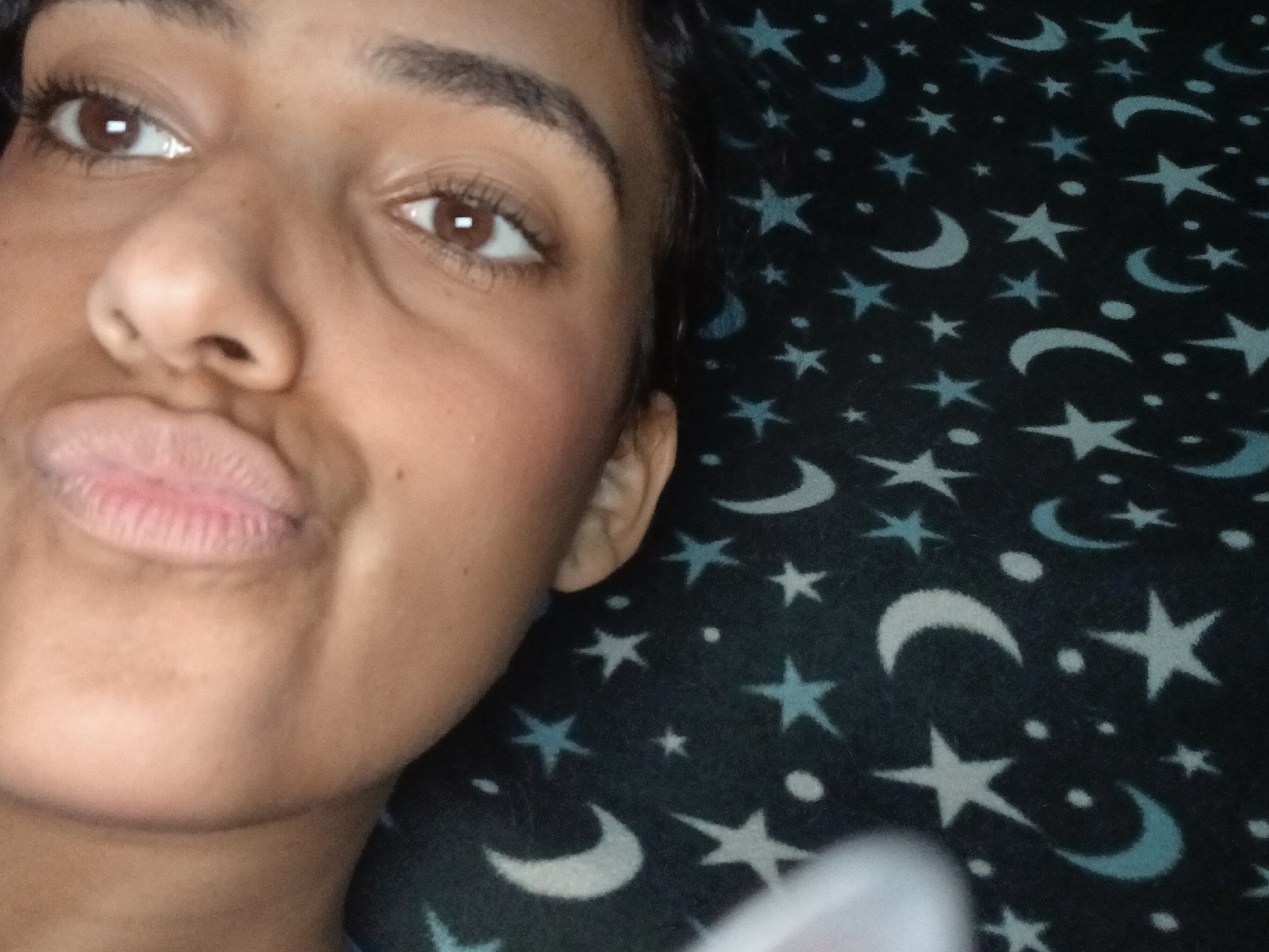 A close-up selfie of a person with brown eyes and puckered lips. They have brown skin and dark eyebrows. The background is a dark fabric with a pattern of light blue stars and crescent moons. The photo appears to be taken indoors.