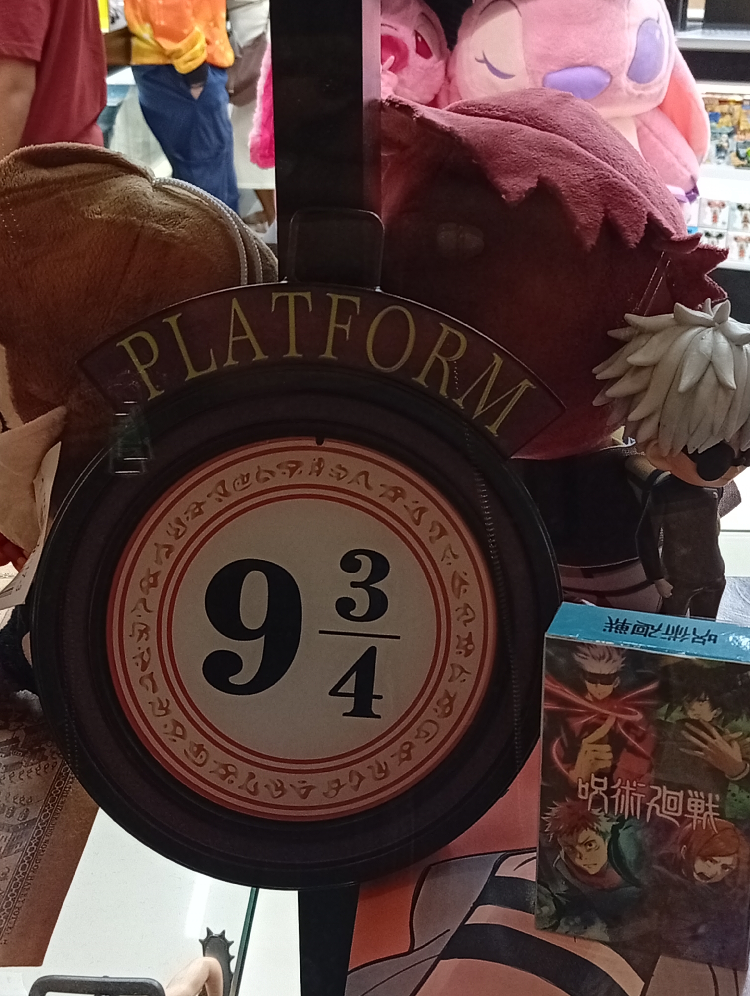 A close-up shot focuses on a round sign featuring "PLATFORM 9 3/4" in raised lettering, a reference to the Harry Potter franchise. The sign is adorned with decorative patterns and is mounted on a dark circular frame. To the right of the sign, a small anime figurine with white hair and sunglasses stands in front of a blue box with Japanese characters and anime characters. The background is cluttered with plush toys, including pink and red stuffed animals. The scene appears to be an indoor display, possibly in a shop or at a convention, in Pampatar, Venezuela.
