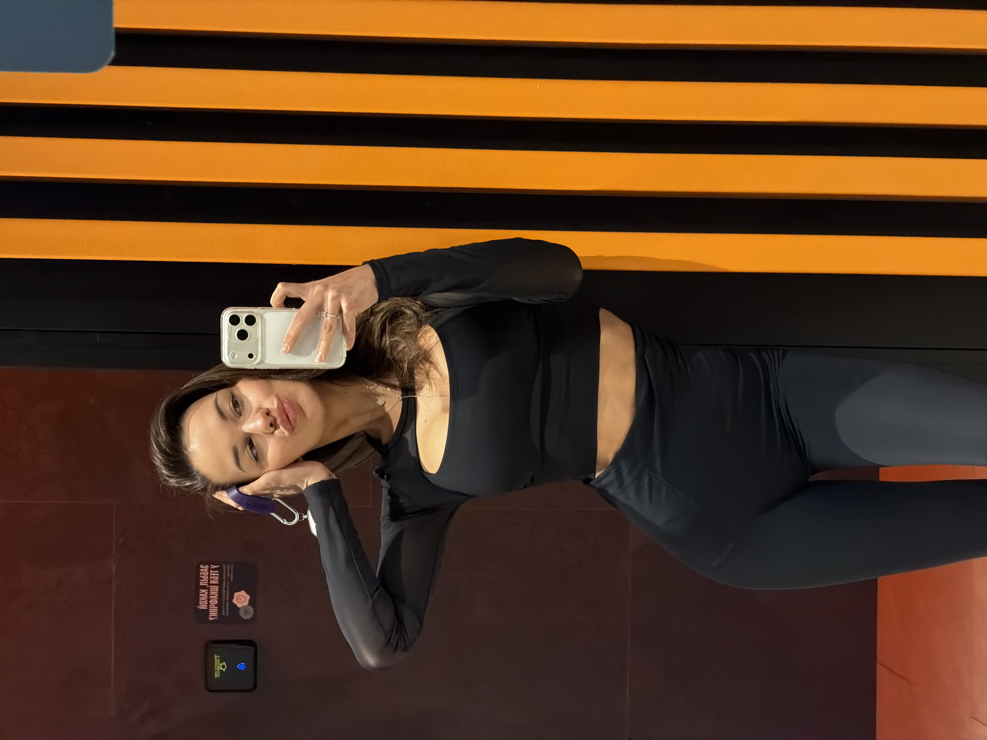 A young woman with long brown hair is taking a selfie using a white smartphone in an indoor location. She is wearing a black long-sleeved crop top and black leggings. Behind her, there is a wall with horizontal orange and black stripes. To her left, there is a brown wall with a sign written in what appears to be Russian and a small electronic device. The scene appears to be in a gym or fitness center, and based on the presence of Russian text, it may be located in the city of Krasnoyarsk, Russia.