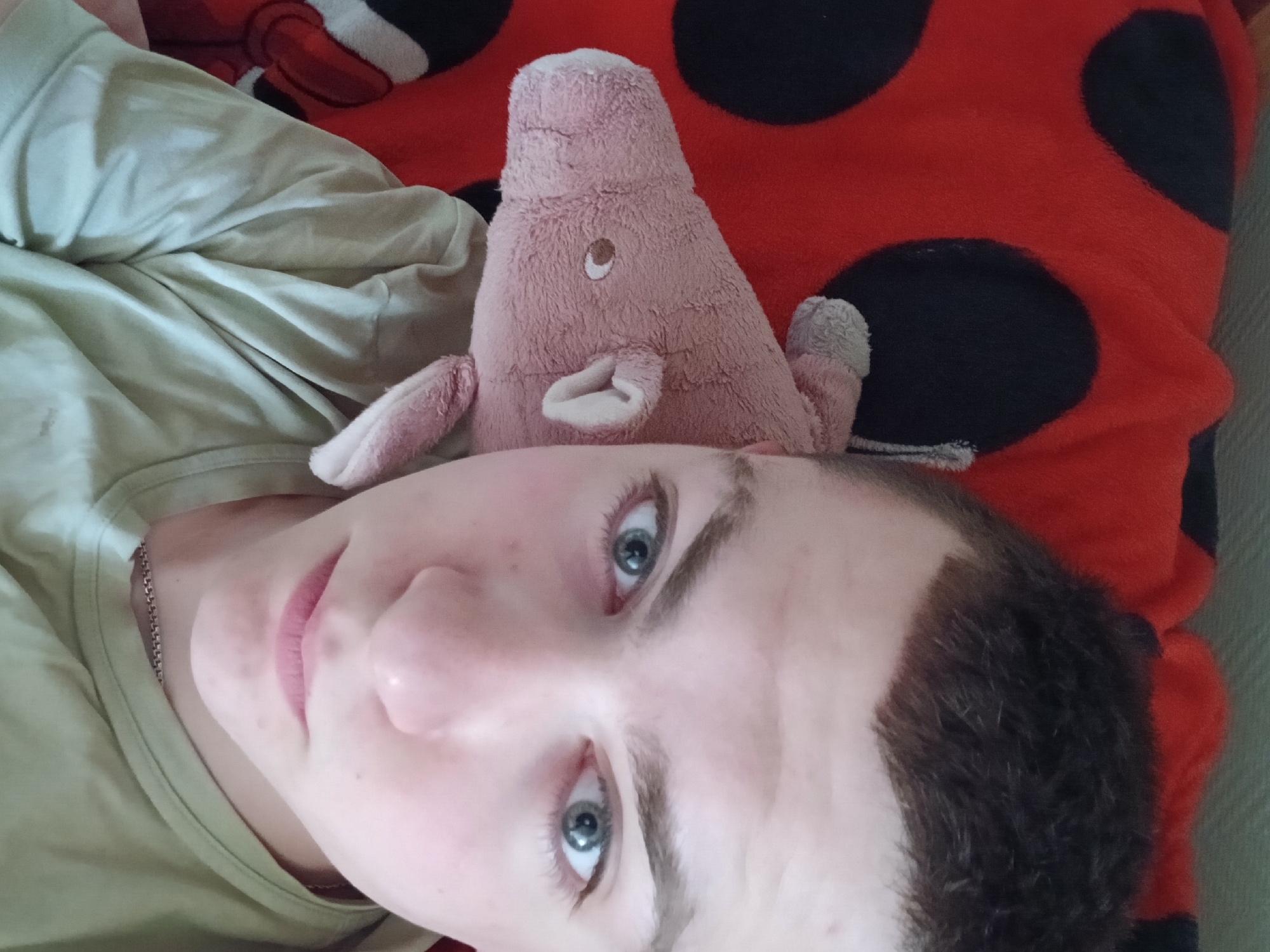 A photograph depicts a person with fair skin, short dark brown hair, and light blue eyes, lying down next to a plush pink pig toy. The person wears a light-colored shirt and a thin silver chain around their neck. The pig toy rests on a red and black polka-dotted blanket. The image was taken from above, with the person's face angled toward the camera. The location is within Chaikovsky, Russia.