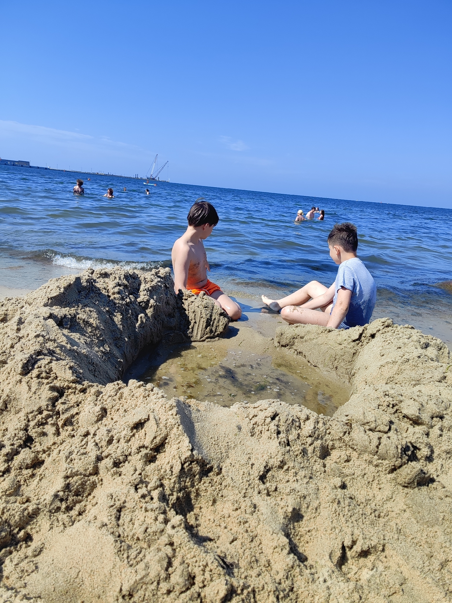 Two young boys are sitting on a sandy beach next to a large, partially constructed sandcastle. The boy on the left is wearing orange shorts and is shirtless. He is sitting with his legs crossed and facing away from the camera. The boy on the right is wearing a blue t-shirt and shorts. He is sitting cross-legged and facing away from the camera, looking out at the water.
The sandcastle has a large, shallow pool of water in the center. The ocean is calm with small waves lapping at the shore. In the background, several other people are swimming and wading in the water. The sky is clear and blue, indicating a sunny day. In the far distance, there appear to be some industrial structures and cranes, suggesting a port or industrial area. The scene depicts a leisurely day at the beach, with the boys engaged in building a sandcastle. The overall mood is relaxed and summery.