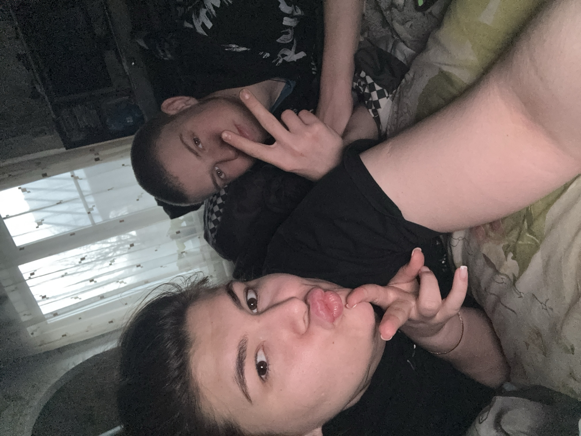Two people are lying on a bed, presumably in a room with a window. The window has multiple panes and appears to be closed. The person at the top of the image is making a peace sign with their fingers. The person at the bottom of the image is pursing their lips and holding a finger near them. The setting, Щучье Озеро, Russia, is not discernible from the provided image.