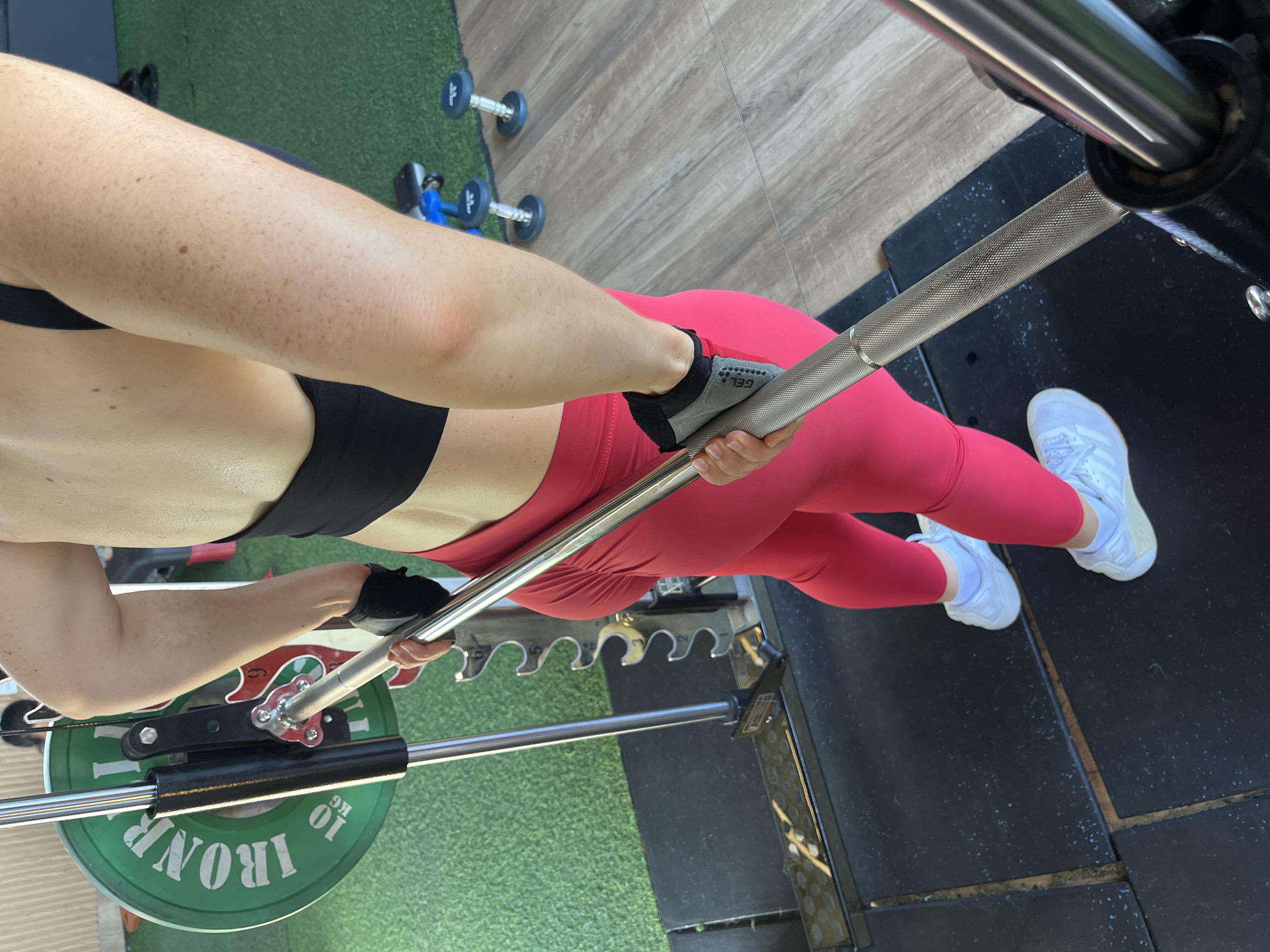 A person is positioned in a gym, gripping a barbell. The person is wearing red leggings, a black sports bra, and white sneakers. Black weightlifting gloves are on their hands. A green weight plate with "IRON" and "10 KG" is visible on the barbell. The gym floor is a combination of black rubber mats and wood-grain flooring. In the background, dumbbells are visible. The image is cropped to focus on the person's upper body and legs, with the barbell extending diagonally across the frame. The setting appears to be an indoor fitness facility. There is no identifiable information linking this scene to Ham Thang Ward, Vietnam.