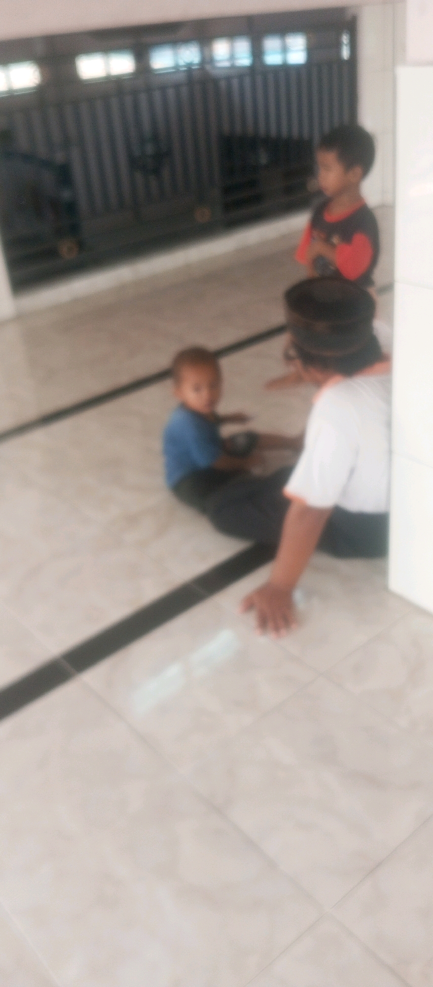 Three individuals, two children and one adult, are situated on a tiled floor, likely indoors. One child, wearing a blue shirt and dark pants, is seated and looking towards the viewer. Another child, positioned slightly behind and to the right, wears a black shirt with red sleeves and appears to be looking away. An adult, wearing a white shirt with orange trim and a black cap, is seated and facing the first child, with their arm extended and hand resting on the floor. The background features a dark, slatted structure with windows, possibly a fence or gate, and a white wall to the right. The scene is blurry, suggesting low resolution or motion blur. The location is identified as Kediri City, Indonesia, though the specific context within the city is not discernible from the image alone.
