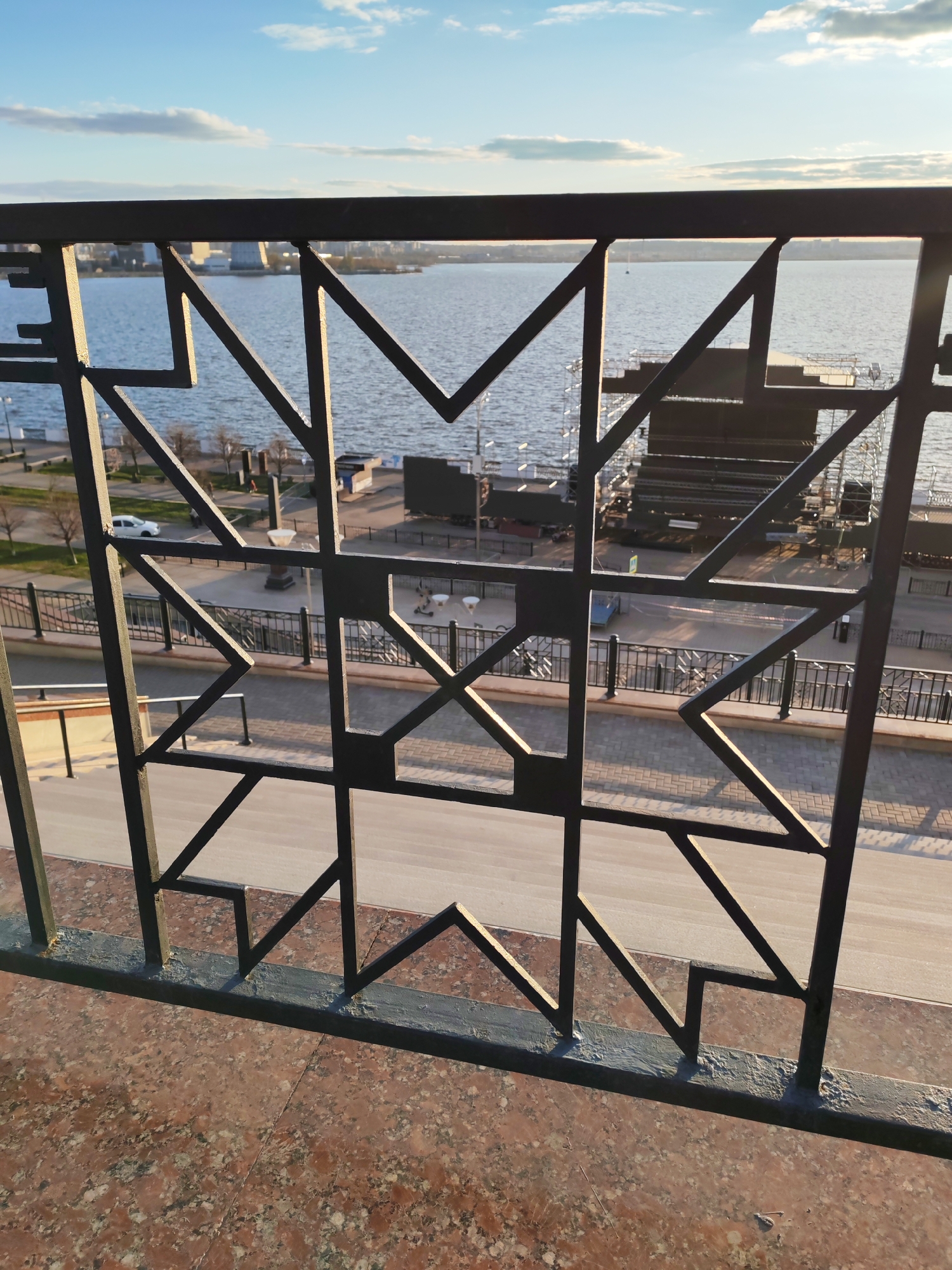 A ground-level, medium shot shows a decorative, black metal railing, behind which is an urban waterfront scene with a river or large body of water, possibly the Izhevsk Pond in Shaberdino, Russia. The railing's design consists of geometric shapes, primarily triangles and squares, creating a star-like pattern. Beyond the railing, a paved walkway is visible, along with street furniture, including benches, lamp posts, and low fences. The background shows structures that appear to be related to stage construction. The sky is blue, with some clouds. A few trees and buildings can be seen in the distance across the water.