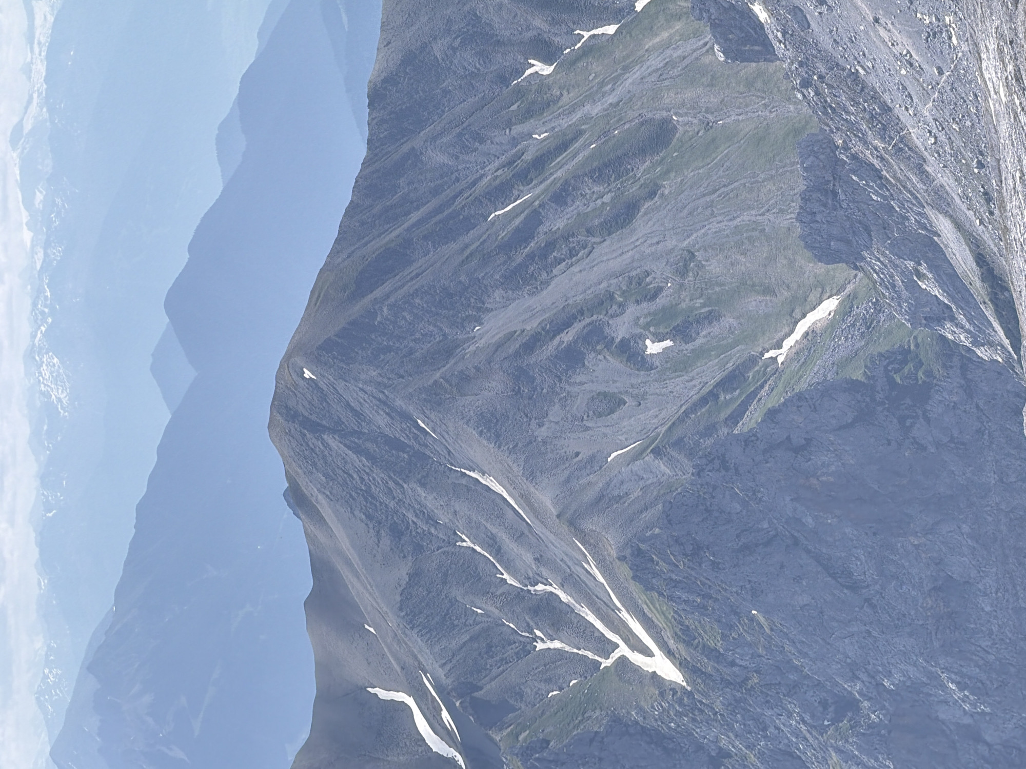 The image displays a mountainous landscape, likely captured from a high vantage point. In the foreground, a rugged mountain slope dominates the frame, characterized by steep, rocky terrain with scattered patches of snow. The texture of the rock is rough and varied, with visible striations and crevices. Green vegetation, appearing sparse, can be seen clinging to some of the less steep inclines. Several white patches of snow are visible, following the contours of the mountain and suggesting meltwater channels.
In the mid-ground, another prominent mountain peak rises, its slopes similarly textured and marked by streaks of snow. This peak exhibits a sharper, more defined summit than the foreground mountain. Beyond this, layers of receding mountain ranges fade into the distance, rendered in progressively lighter shades of blue due to atmospheric perspective. The furthest mountains appear as soft, hazy silhouettes against a bright, overcast sky.
The scene is devoid of any human presence, structures, or discernible human activity. The environment is purely natural, characterized by geological formations and natural elements.
Considering the geographical context of Garmisch-Partenkirchen, Germany, this image strongly suggests a view of the Bavarian Alps. The prominent peaks, dramatic slopes, and presence of snow are characteristic of this alpine region. While a specific location within Garmisch-Partenkirchen cannot be definitively identified from the image alone, it is representative of the panoramic mountain vistas accessible from the surrounding area, potentially from the Zugspitze or another elevated viewpoint. The overcast sky suggests a cool, possibly humid, weather condition typical of mountain environments.