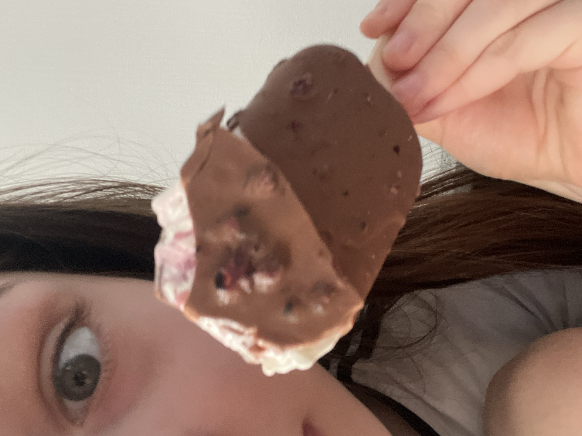 A close-up image features a person's partially visible face and a hand holding an ice cream bar. The left side of the person's face is visible, showing a light skin tone, a left eye with a dark iris and dilated pupil looking directly forward, and a portion of the nose and cheek. Dark brown hair frames the right side of the face. The person is wearing a white garment with a dark band or collar visible on the left side of the neck/shoulder area.
The right hand holds a white stick of an ice cream bar. The ice cream itself has an irregular shape and is largely coated in a dark brown, hardened chocolate layer. This chocolate coating contains small, dark red or purple flecks, possibly pieces of berries or fruit. The coating is broken in places, revealing a white interior that also appears to contain pink or light red streaks or pieces.
The background is an undifferentiated, plain light-colored wall, providing no specific environmental details. The image provides no discernible details to identify a specific geographic location, including the city of Saint Petersburg, Russia.