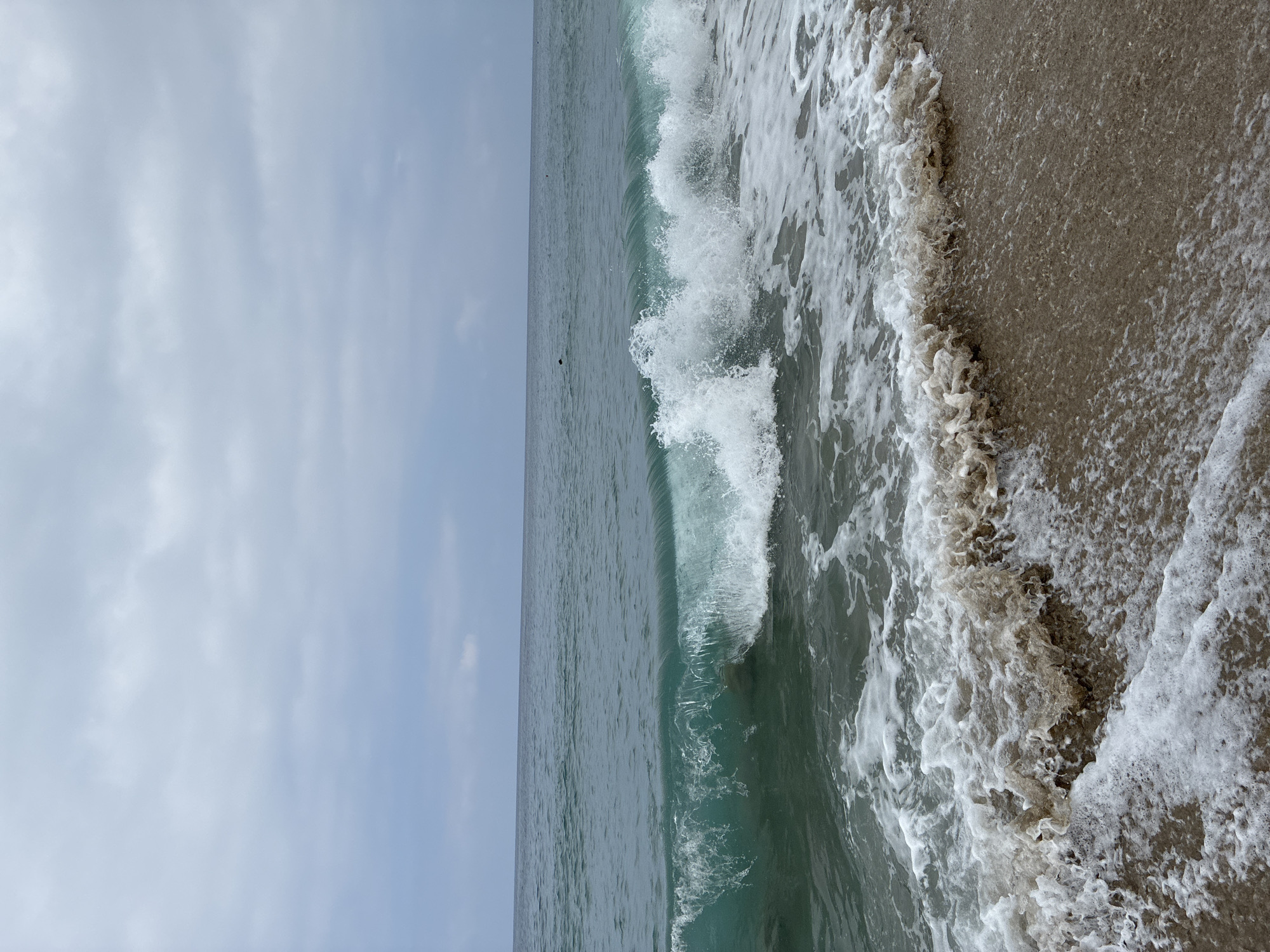 The image depicts a beach scene in Alanya, Turkey, under a cloudy sky. A wave is breaking on the shore, creating white foam and churning water. The ocean water is a greenish-blue color, and the sand is light brown. The horizon line is visible, separating the sea from the sky. The sky is filled with various shades of gray clouds. A few dark specks, potentially people or objects, are visible in the distance on the water. The foreground shows the wet sand with receding foam and bubbles.