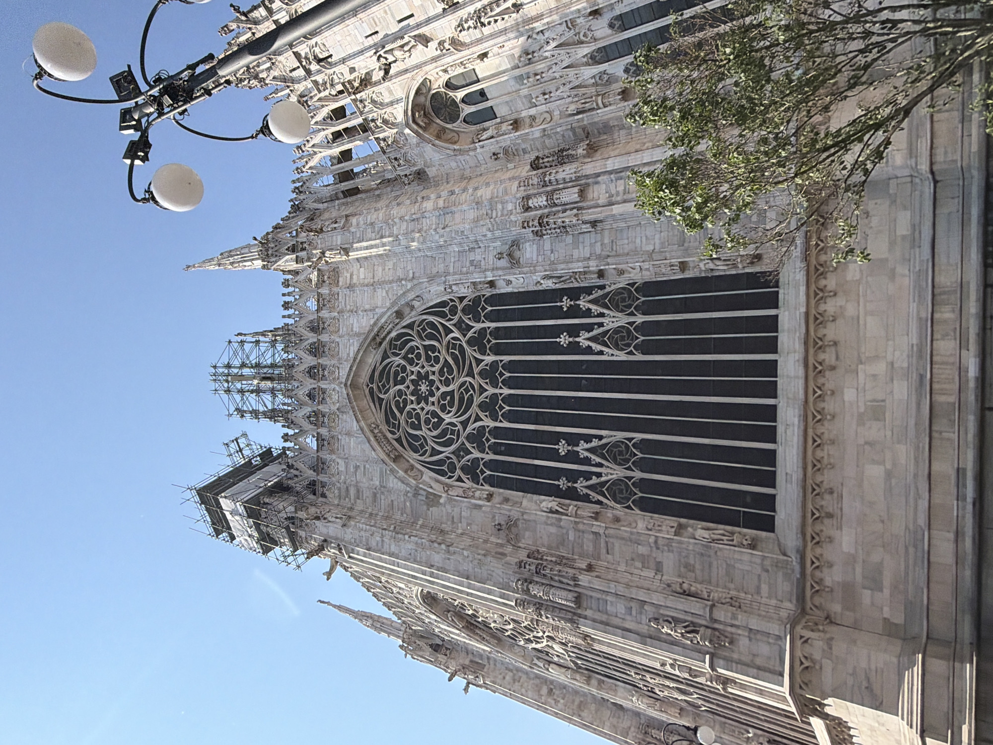 The image shows a low-angle, upward-facing view of the Duomo di Milano, a Gothic cathedral located in Milan, Italy. The facade of the cathedral is constructed with intricate stonework and detailed carvings. Scaffolding covers a section of the building indicating ongoing maintenance or restoration. A portion of a street lamp with rounded light fixtures is visible in the upper left corner of the frame. Bare tree branches appear along the upper right side of the building. The sky in the background is clear and blue.