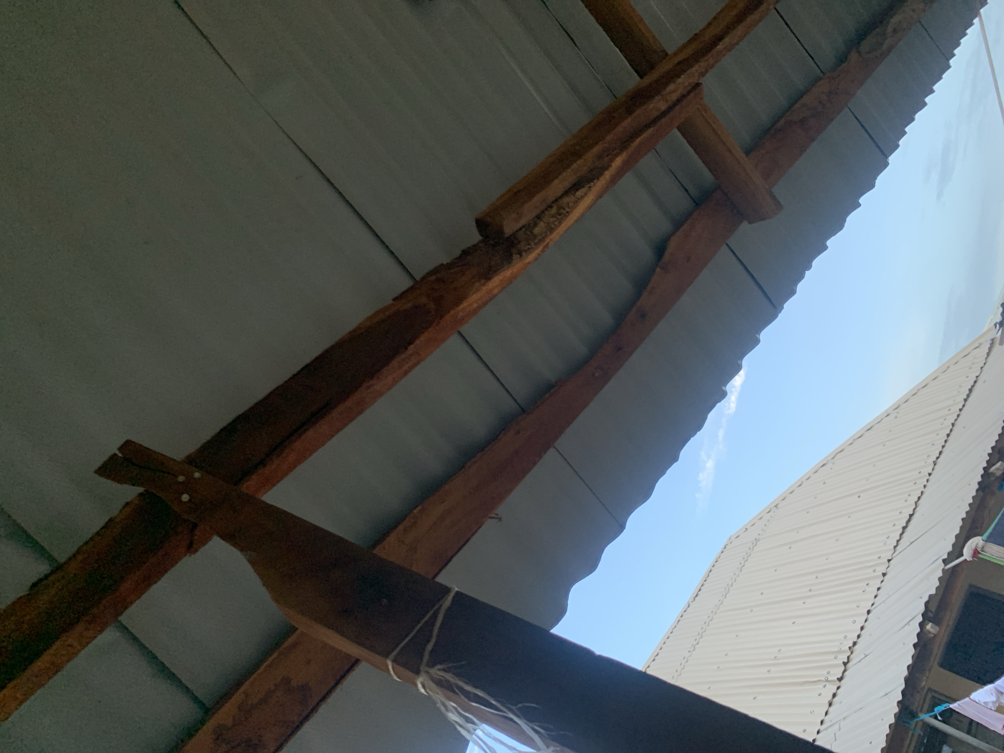 The image provides an upward-angled view of a roof structure, likely an outdoor shelter, porch, or part of a building in Keffi, Nigeria. The primary subject is the underside of a corrugated metal roof, supported by a network of wooden beams. The roofing sheets are a dull grey, running diagonally across the frame, with their characteristic wavy profile clearly visible.
Robust wooden beams, some appearing rough-hewn and others more finished, form the framework beneath the metal sheets. These beams are dark brown, showing natural grain and some signs of wear. One particularly thick, dark wooden beam crosses the lower part of the foreground, with several strands of white string or rope tied around it.
Beyond this immediate structure, a clear, light blue sky with faint white clouds is visible, indicating bright daytime conditions. To the right, another building's roof, also made of light-colored corrugated metal sheets, extends upwards into the frame. This adjacent roof appears brighter, possibly white or very light grey, and is secured with numerous visible rivets or screws. At the far bottom right, part of a wall and what looks like hanging laundry or other items on lines are faintly discernible. The scene depicts functional, simple construction methods typical of the region. No people or visible text are present.