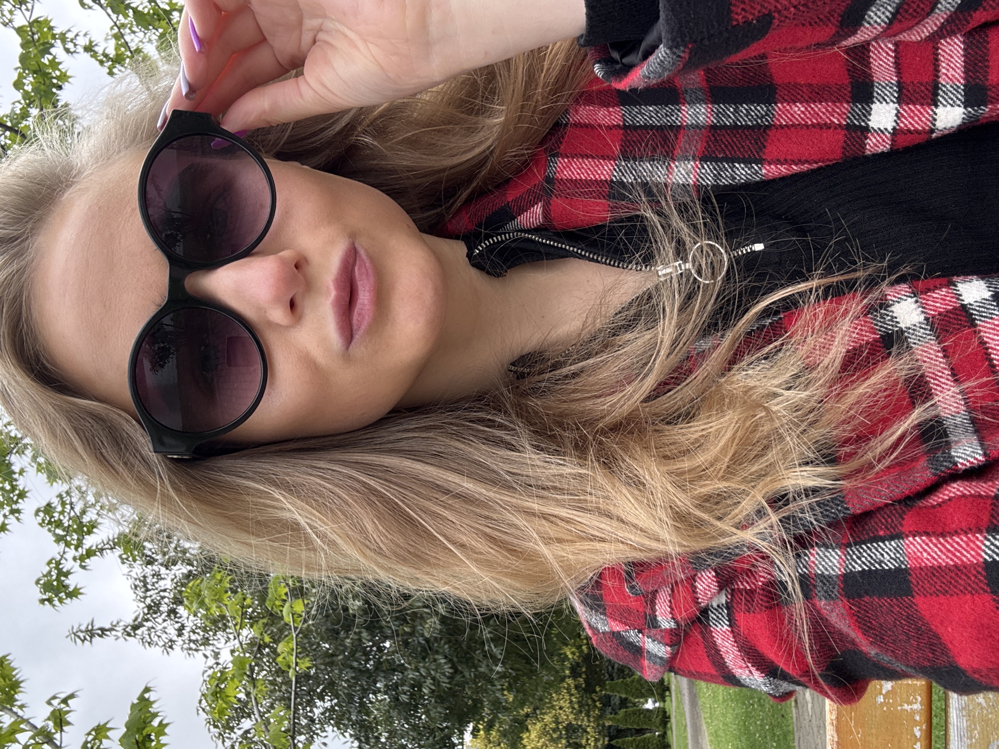 A person with blonde hair is wearing sunglasses and a red and black plaid jacket in an outdoor setting. The person is holding the sunglasses slightly away from their face. The environment includes trees, greenery, and a cloudy sky, suggesting a park or natural area. The location is in the city of Unknown, Unknown.