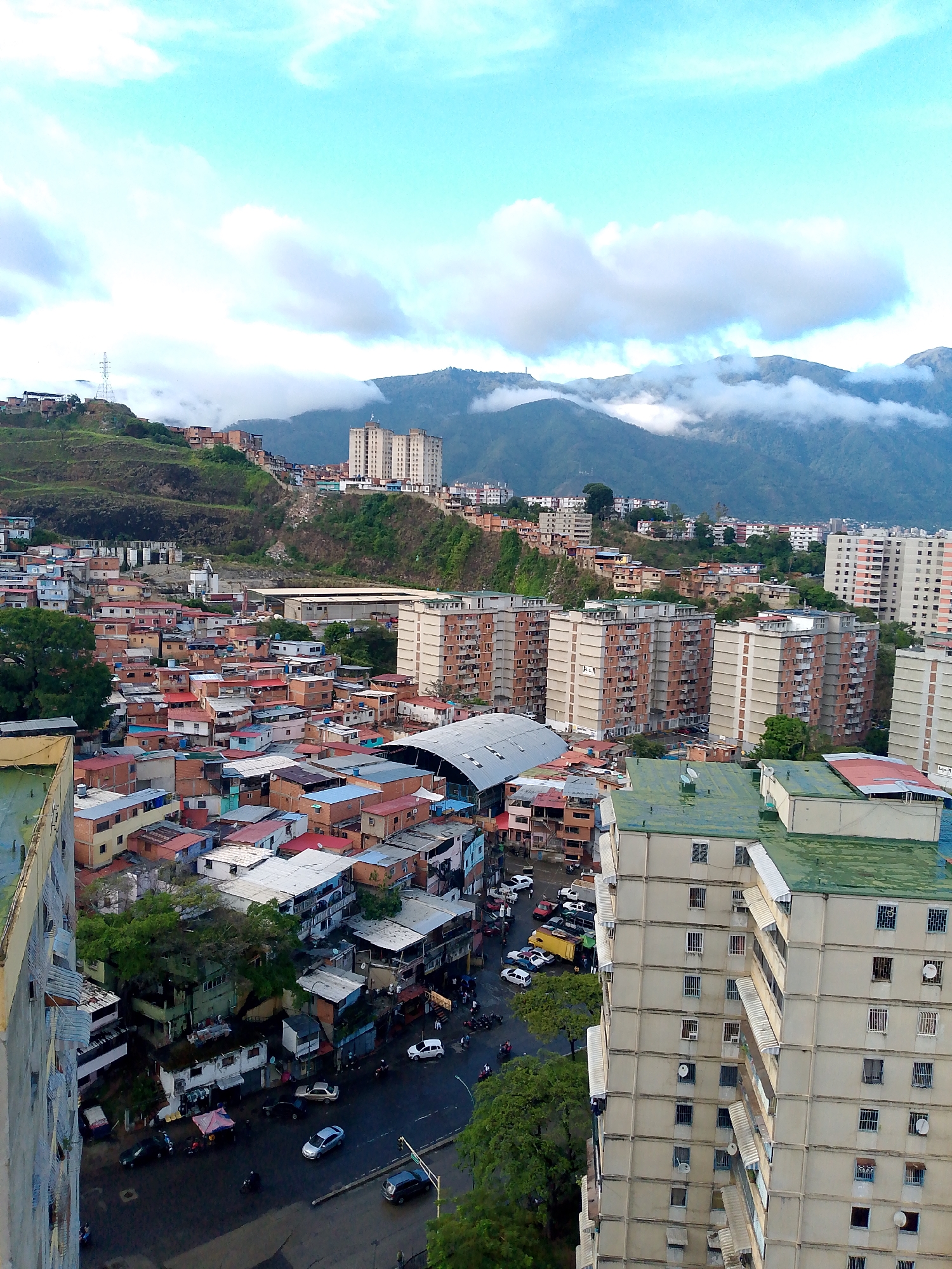 The image depicts a sprawling urban landscape in Caracas, Venezuela, characterized by densely packed residential buildings and a backdrop of lush, cloud-shrouded mountains. The foreground is dominated by the side of a large, light-colored apartment building with numerous windows, some equipped with awnings and protective grilles. Below and to the left, a jumble of smaller, multi-colored houses with red and blue roofs create a stark contrast to the towering apartment blocks. Streets are visible, with several vehicles including cars and motorcycles, and a few pedestrians can be seen. The scene conveys a sense of everyday urban life in a hilly environment, with the natural landscape playing a significant role in the city's topography. The presence of satellite dishes on rooftops is a common sight in urban areas. The lighting suggests it is daytime, possibly late afternoon or early morning, with soft light and some cloud cover.