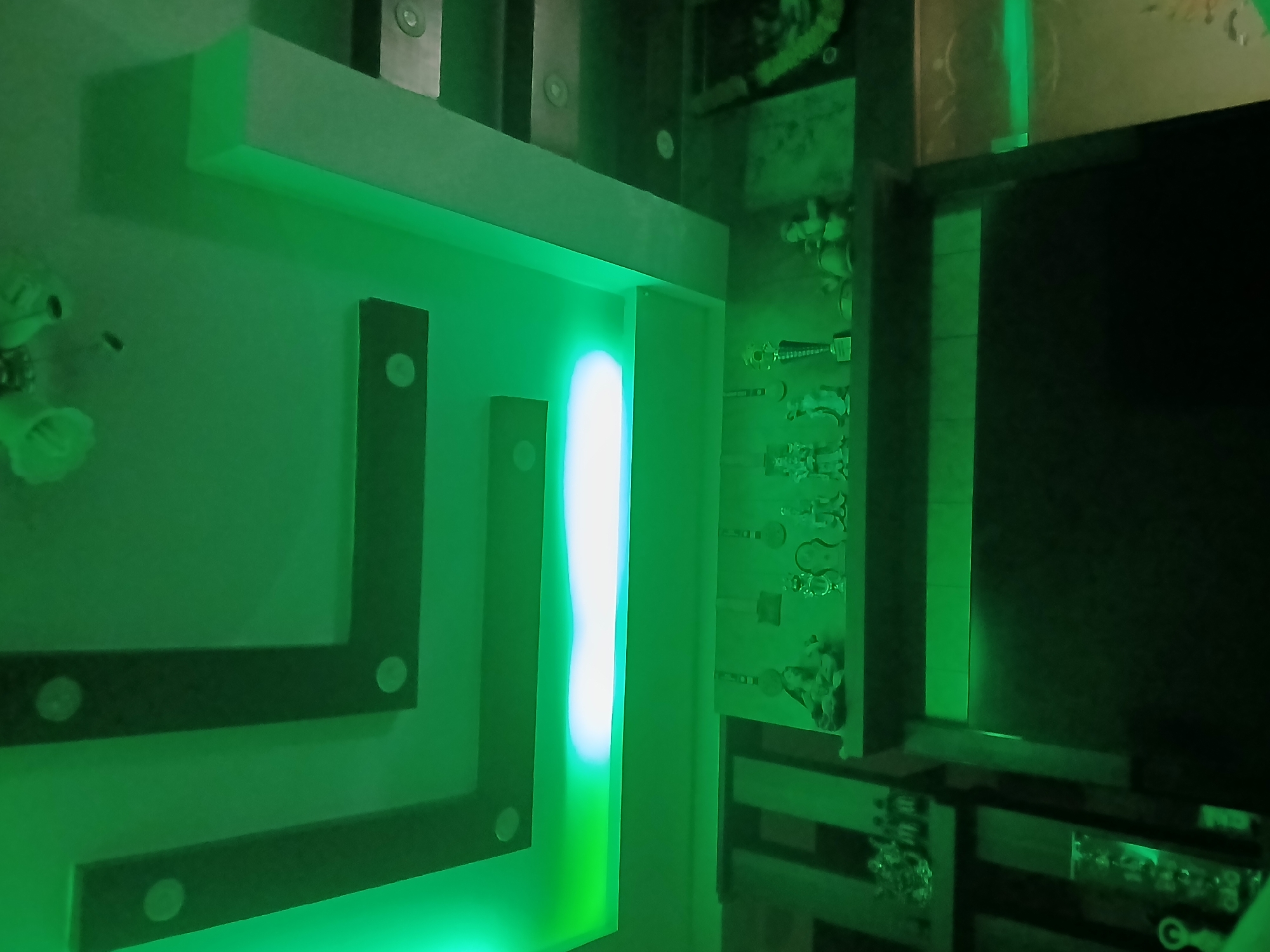 This media file captures a contemporary interior space illuminated predominantly by green light, suggesting an evening or night setting. The scene is dominated by a wall featuring modern, geometric decor.
On the left side, there's a light-colored, possibly white, horizontal shelf or panel extending from the upper wall, casting a green glow downwards onto a light green wall surface. Below this, dark, rectangular panels are mounted on the wall in an "L" shape, adorned with small, circular, light-colored inserts.
A prominent vertical light strip emits a bright white and blueish glow, contrasting with the overall green ambient lighting, and is positioned between two dark vertical panels. To the right, another section of the wall features multiple small, decorative figurines or statues arranged on shelves, all bathed in the green light, making their details somewhat obscured but appearing to be human or mythical figures. Some resemble trophies or awards.
Further to the right, a large, dark rectangular panel, possibly a television screen or a built-in display, occupies a significant portion of the frame. Below this panel, dark shelves or cabinets are visible, with some reflective surfaces and hints of other decorative items or components. The upper part of the image shows more architectural details and embedded circular lights, consistent with modern false ceiling or wall designs.
The overall atmosphere is cool and modern, with the green lighting creating a distinct mood. There is no activity or visible text in the image. The location is contextualized as Bengaluru, India, but no specific visual cues within the image confirm this.