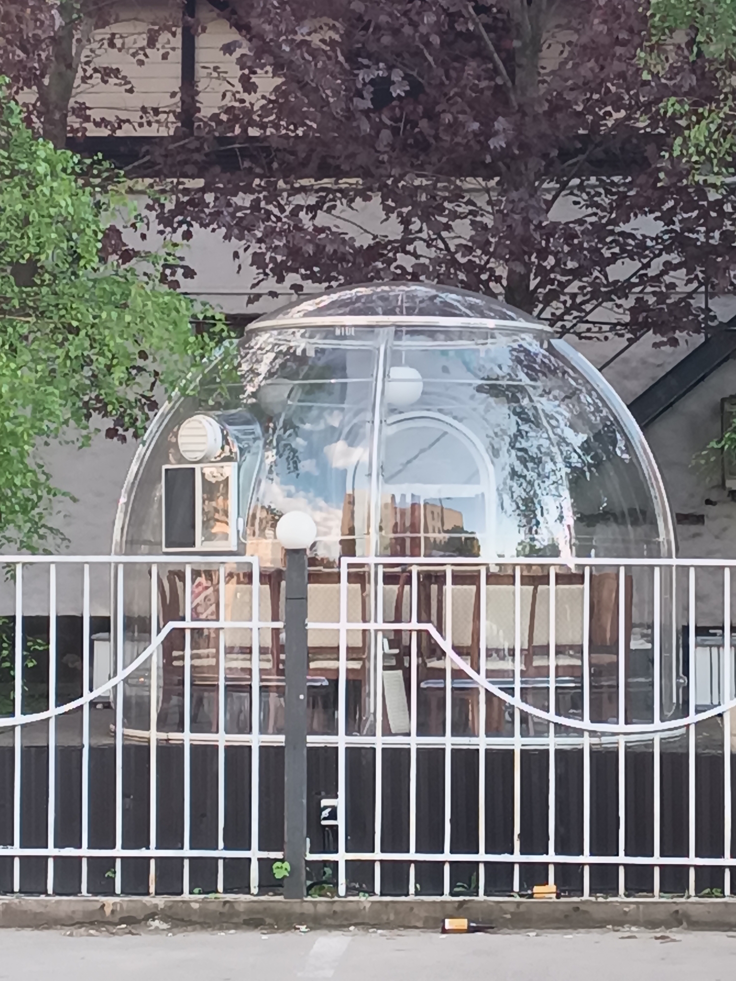 The image depicts a transparent dome-shaped structure, possibly a dining enclosure, located behind a white metal fence. Inside the dome, several chairs and tables are visible, suggesting a dining or seating area. A light post is positioned in front of the dome, partially obscured by the fence. The background includes trees with green and burgundy leaves and a building with visible architectural details. The scene is set on a street or pathway, with the bottom part of the fence and some trash visible near the ground. The location is described as being in the city of Unknown, Unknown.