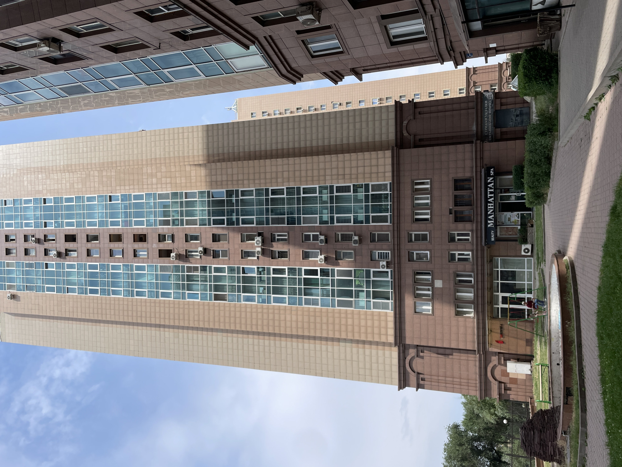 The image shows a view of a residential high-rise building complex in Almaty, Kazakhstan. The primary building in the foreground is a tall, modern structure with a beige facade and a central section of blue-tinted windows. Below the windowed section, the building has a darker brown, stone-clad base with multiple shops and entrances at ground level. One shop is labeled "MANHATTAN SPA." To the left of the main building, there is a children's play area with swings and a dry fountain. To the right, another similar high-rise building is partially visible. The sky above is blue with scattered clouds. The overall scene depicts an urban residential environment with commercial spaces on the lower floors.